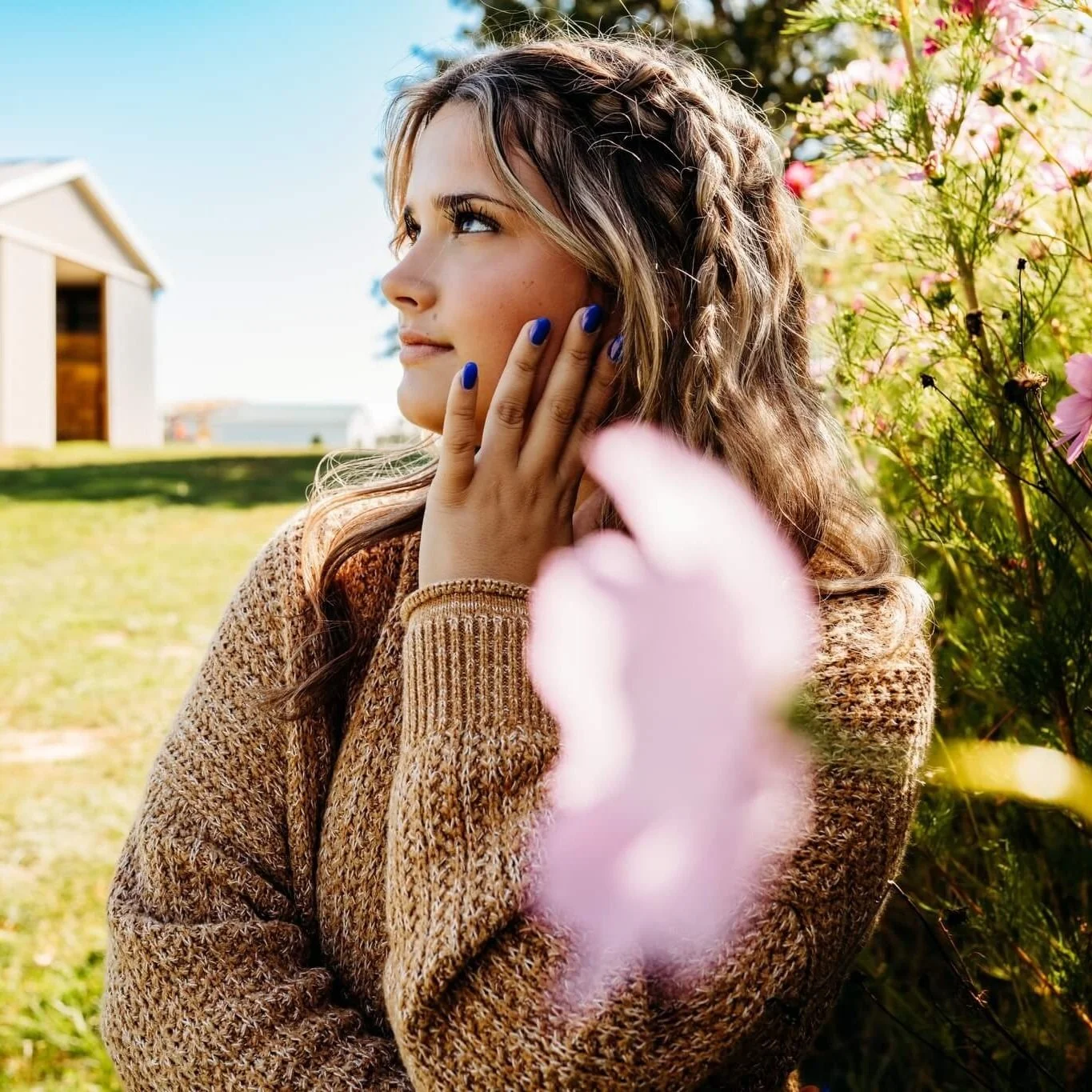 Young woman with wavy hair and blue nail polish, wearing a brown sweater, standing outdoors near pink flowers, looking to the side with her hand on her face.