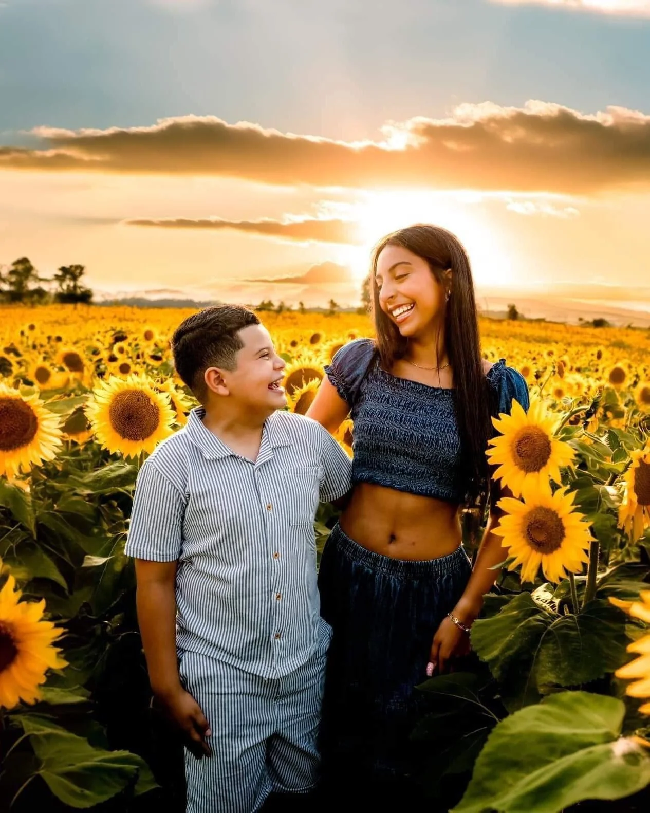 A woman and a young boy standing in a sunflower field at sunset, smiling at each other.