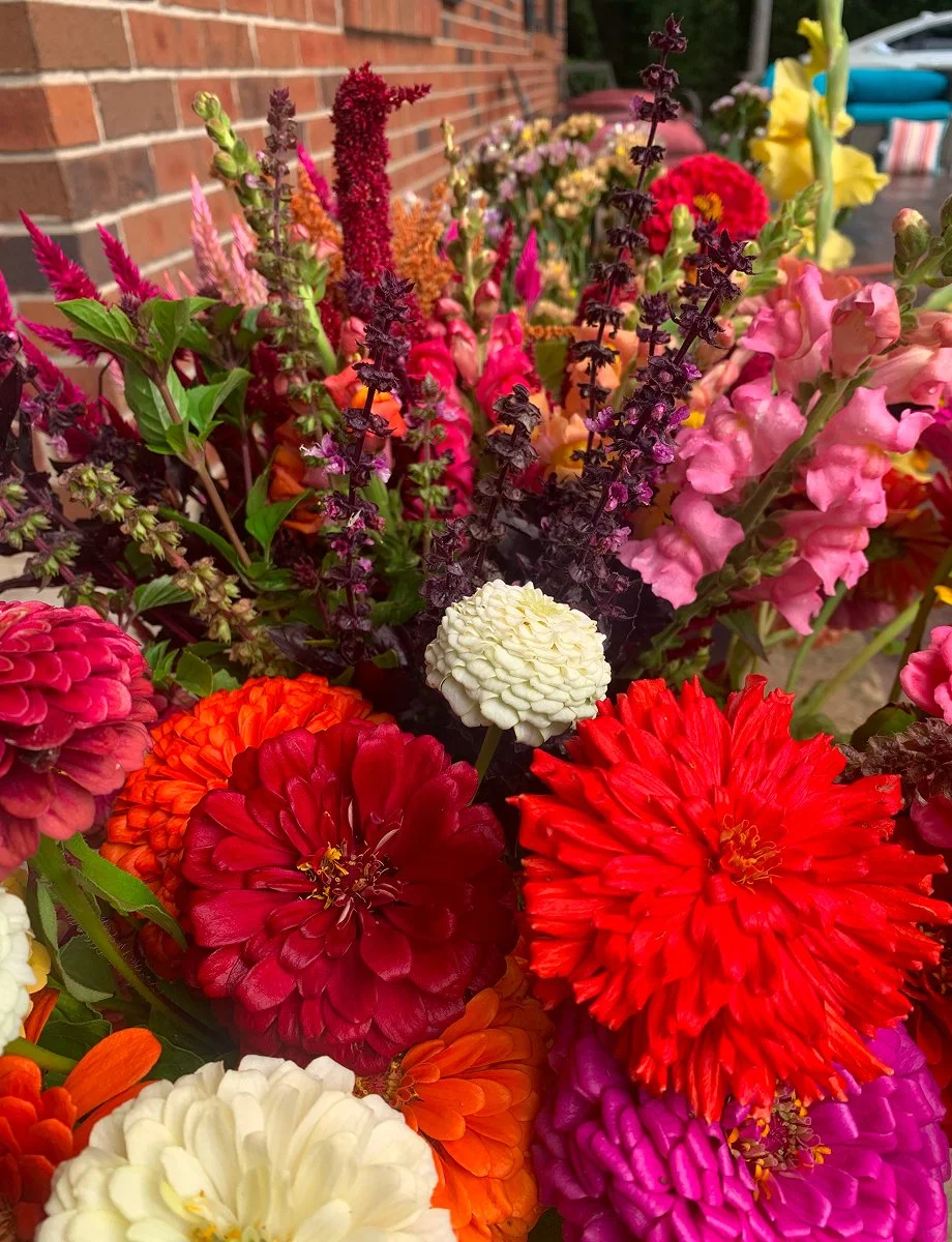 Colorful bouquet of freshly cut summer flowers, including zinnias, snapdragons, and basil, displayed in front of a brick wall.