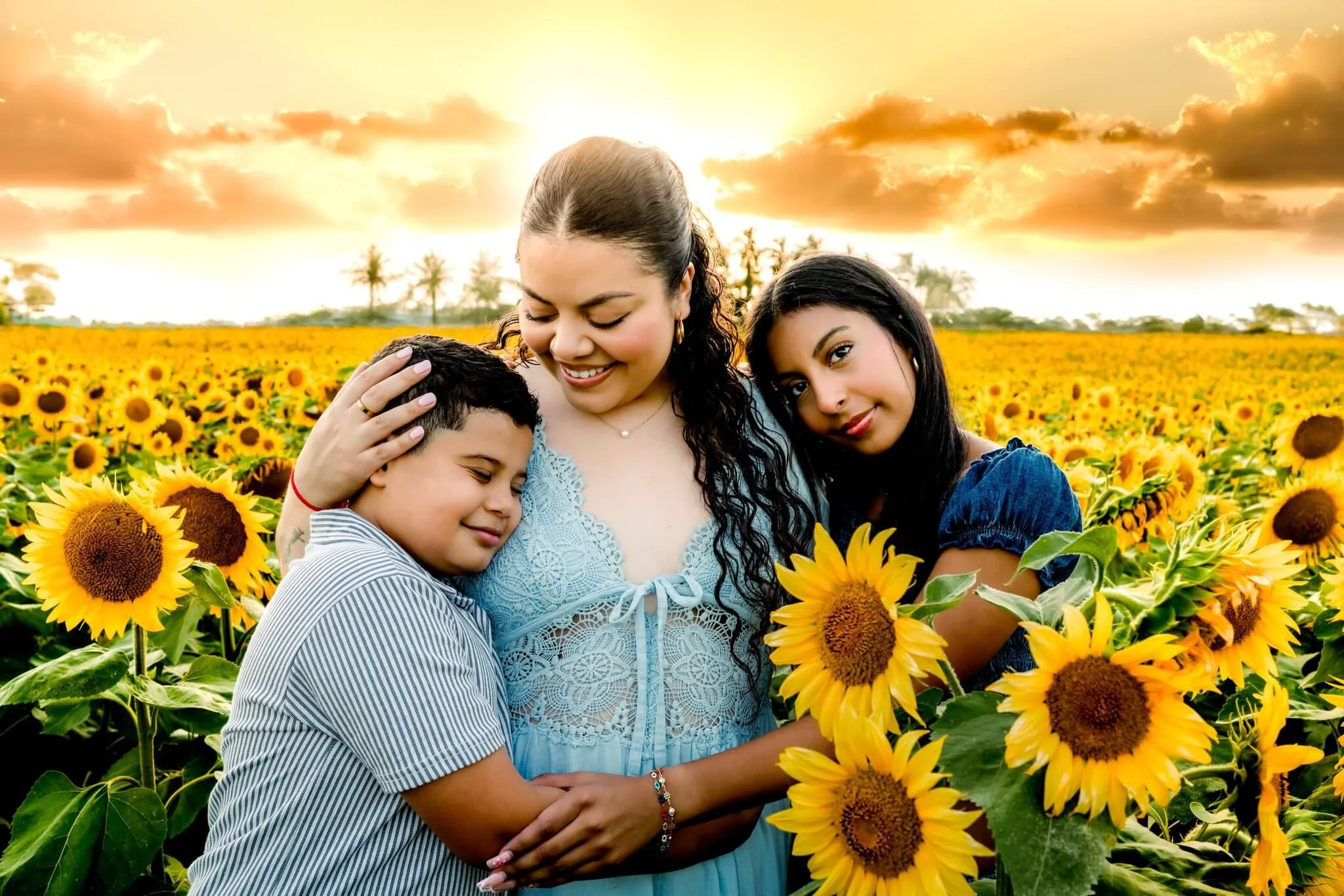 Three women standing together in a sunflower field during sunset, embracing and smiling.