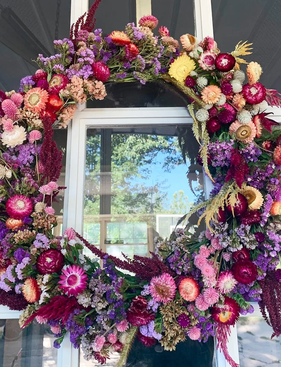 Large handmade floral wreath featuring dried blooms in purple, pink, red, and cream tones, hanging on a window.