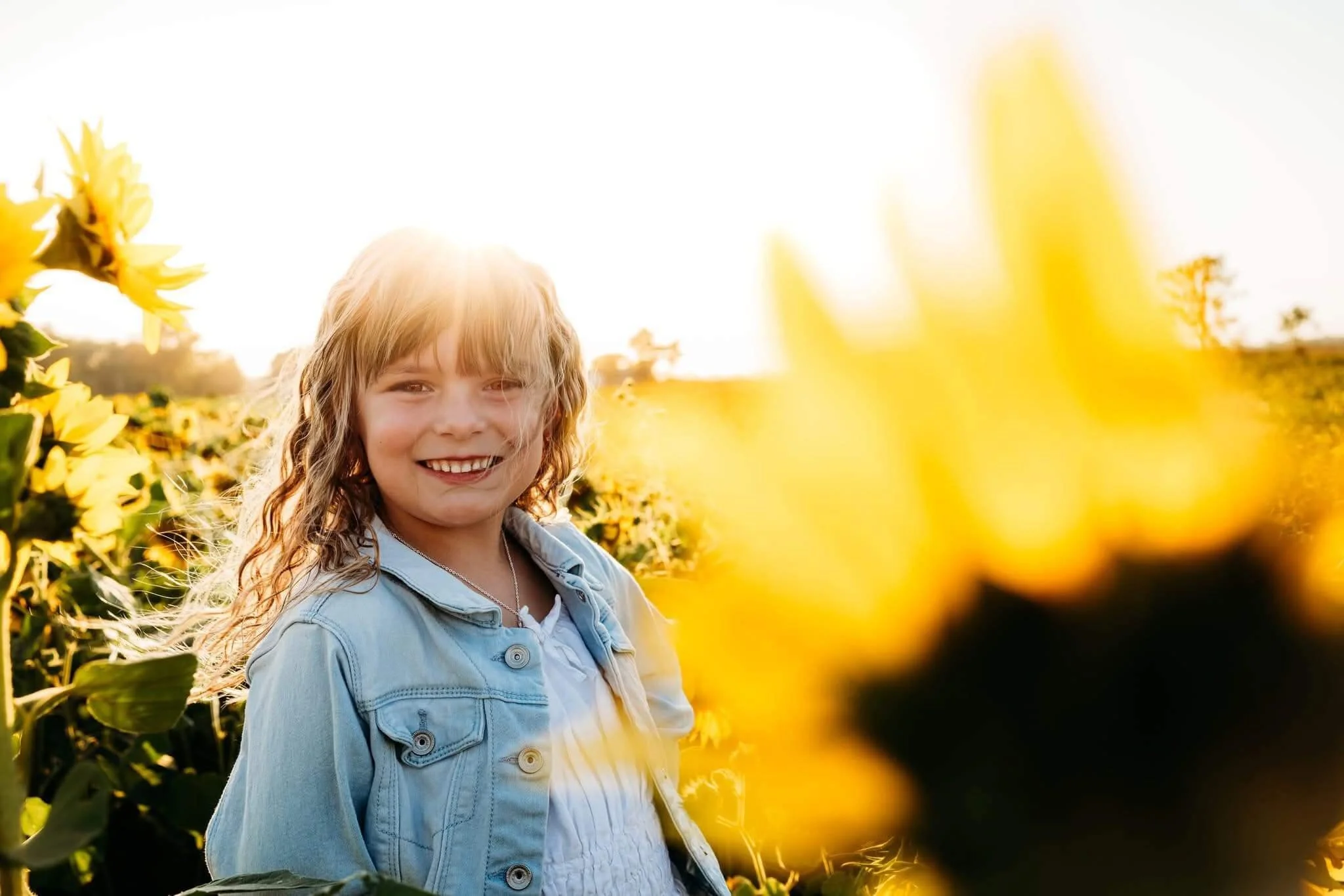 Young girl with curly blonde hair smiling in a sunflower field during sunset, wearing a light blue denim jacket and white top.
