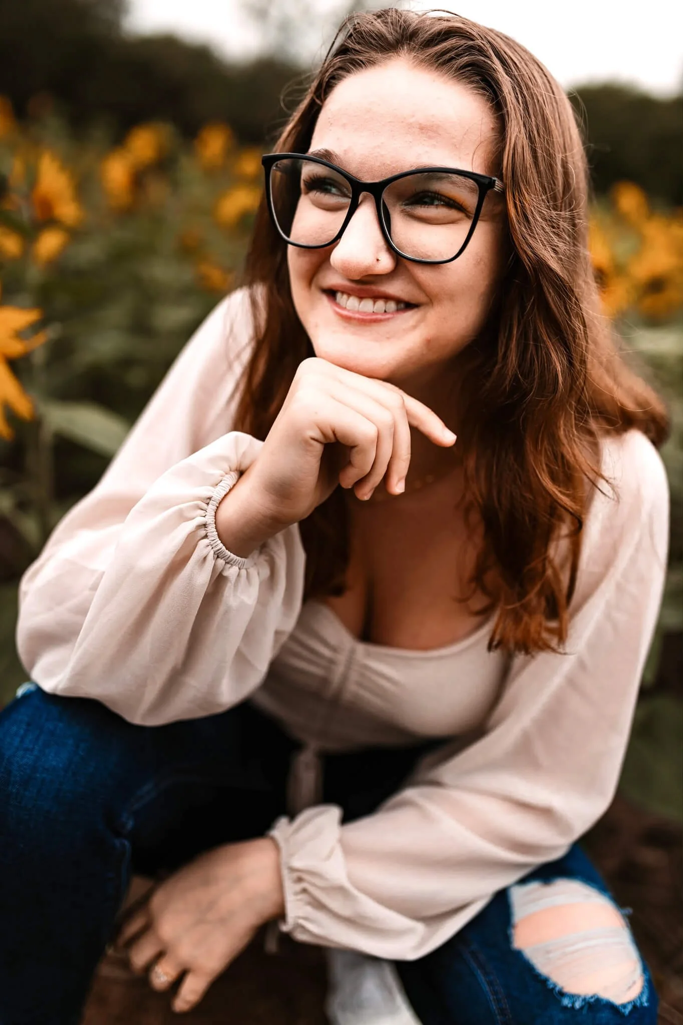 A woman with long red hair, glasses, and a beige blouse, smiling and sitting outdoors near yellow sunflowers.
