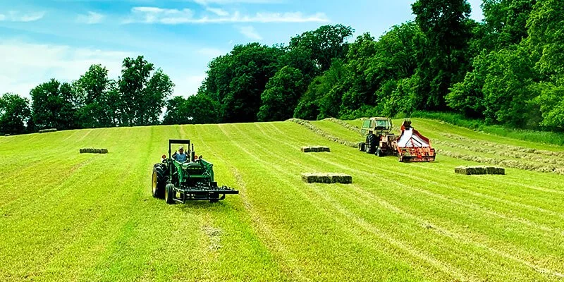 Two tractors in a large grassy farm field harvesting hay, with stacks of hay bales and a tree line in the background under a partly cloudy sky.