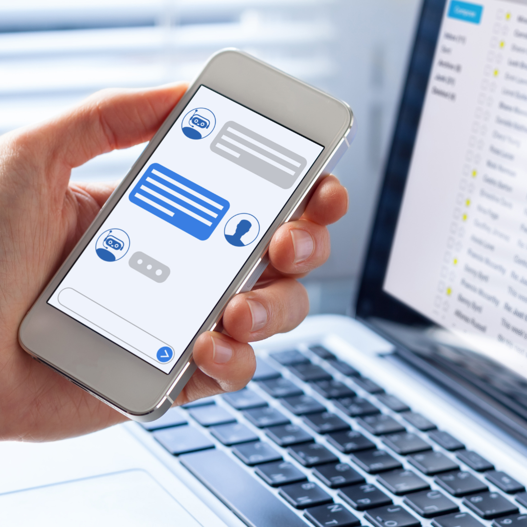 Person holding a smartphone displaying a chatbot conversation with robot and human icons, in front of a laptop showing an email inbox.