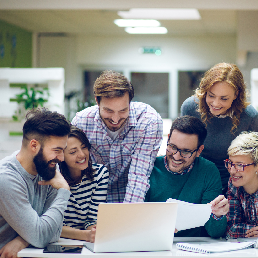 Group of six people gathered around a laptop, smiling and looking at the screen, in a brightly lit indoor office or workspace.