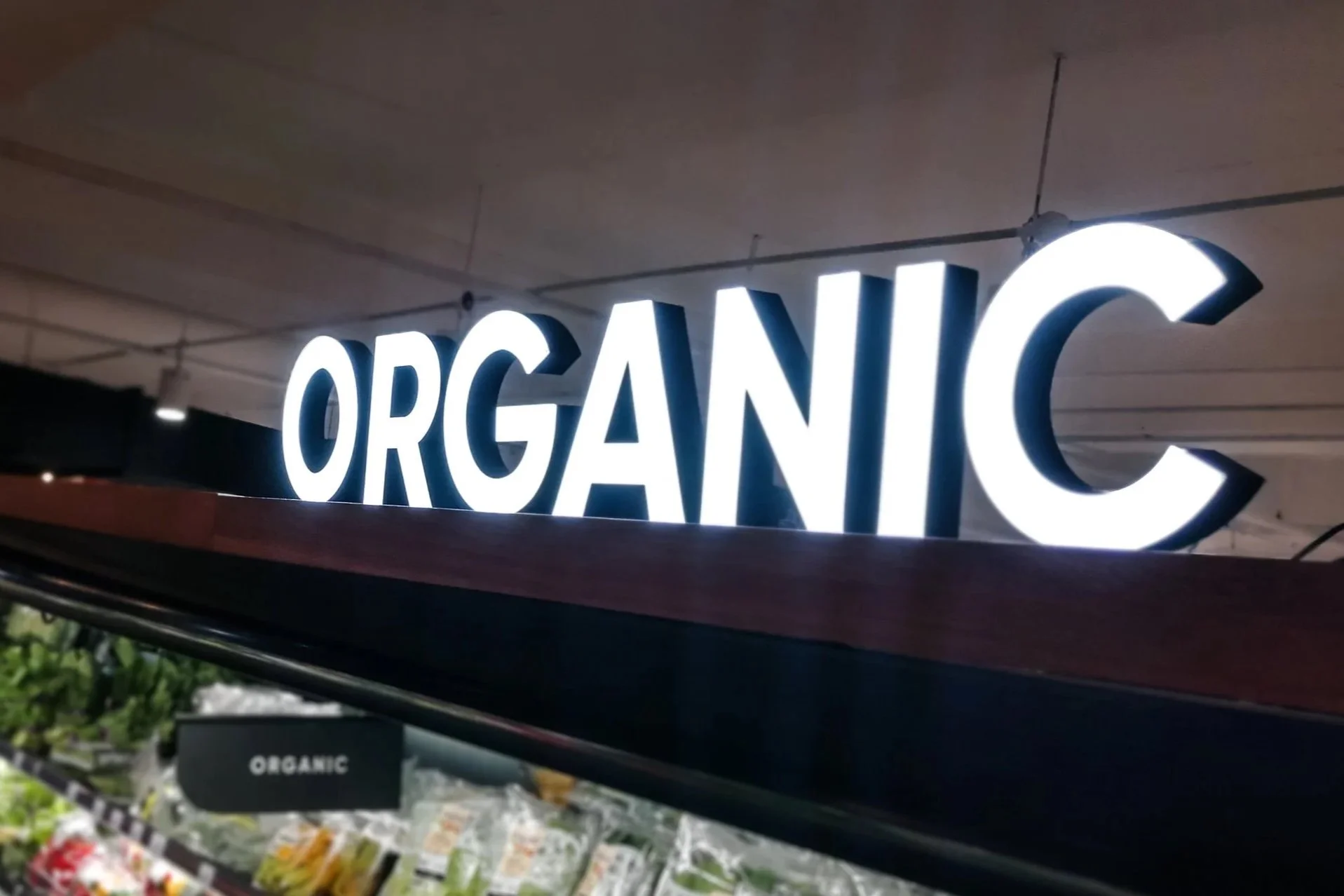 An outdoor sign with placeholder text for custom design and logo, mounted on a building with trees and sunlight in the background.