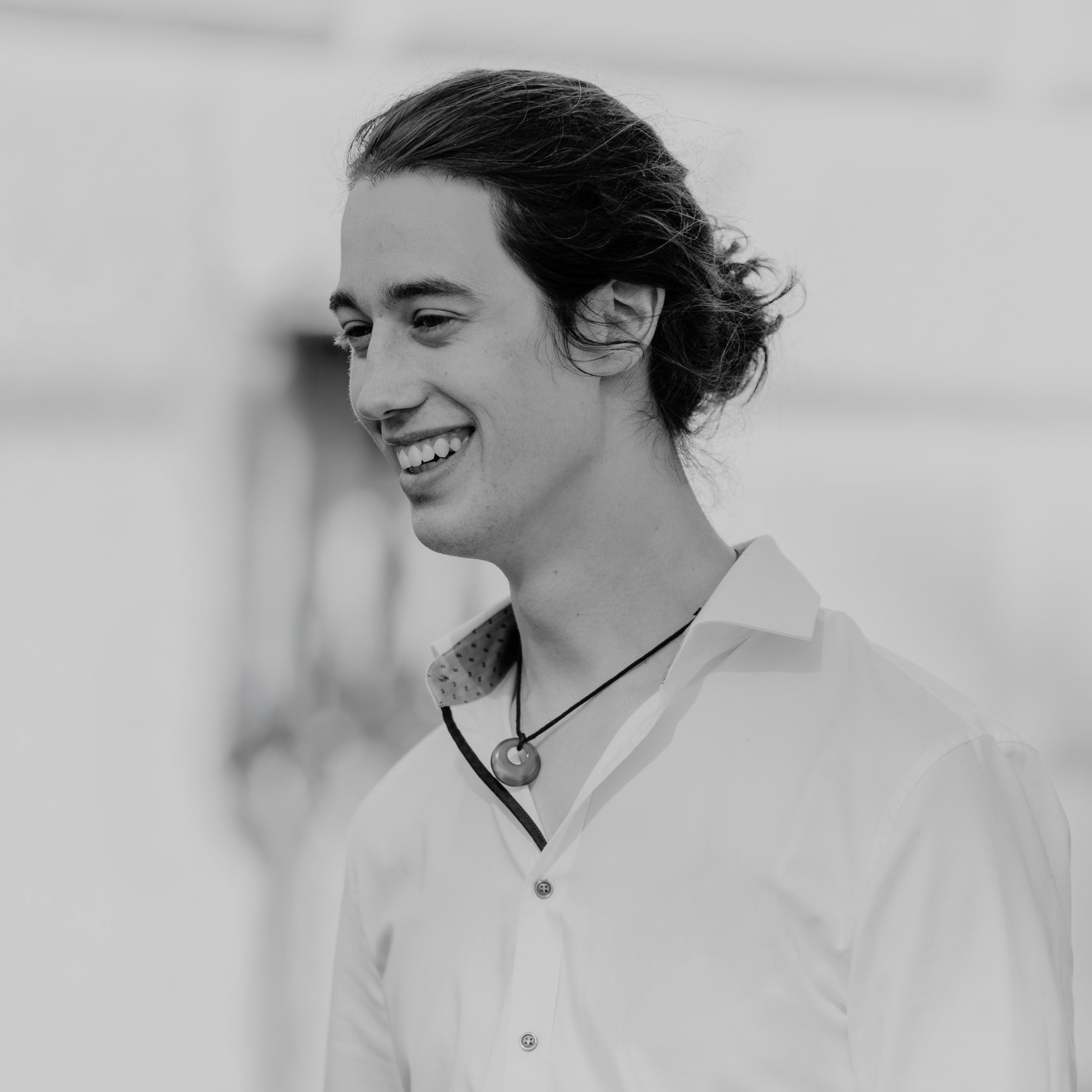 Black and white photo of a young man with long, dark hair, smiling and wearing a collared shirt with an open neckline and a necklace with a circular pendant.