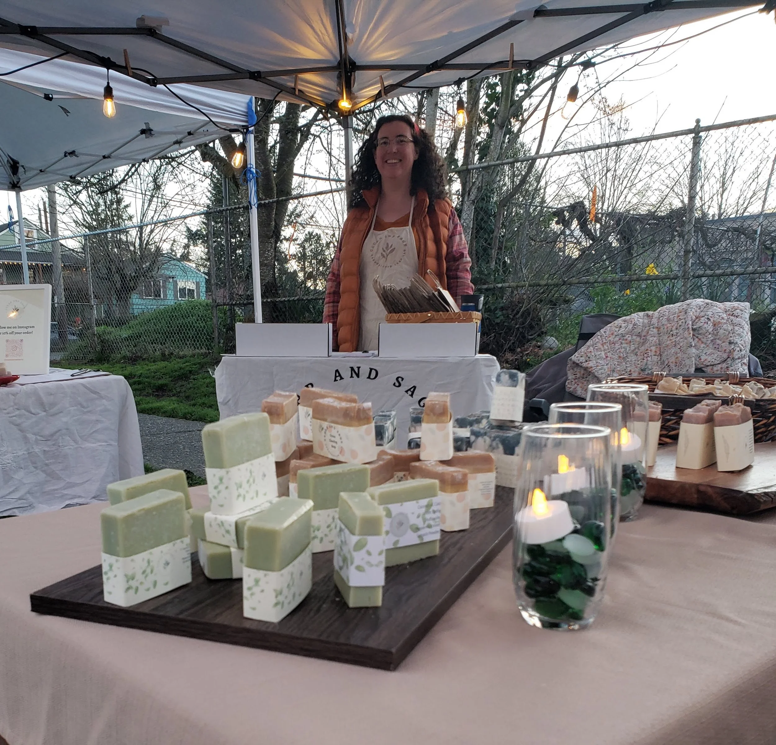A woman standing behind a table with handmade soap products at an outdoor market stall during dusk. The table displays green and beige soap bars, a glass filled with green and white stones, and other soap items. The woman is smiling and wearing glasses, an apron, and an orange puffer vest. String lights hang overhead, and a fence with trees and houses is visible in the background.