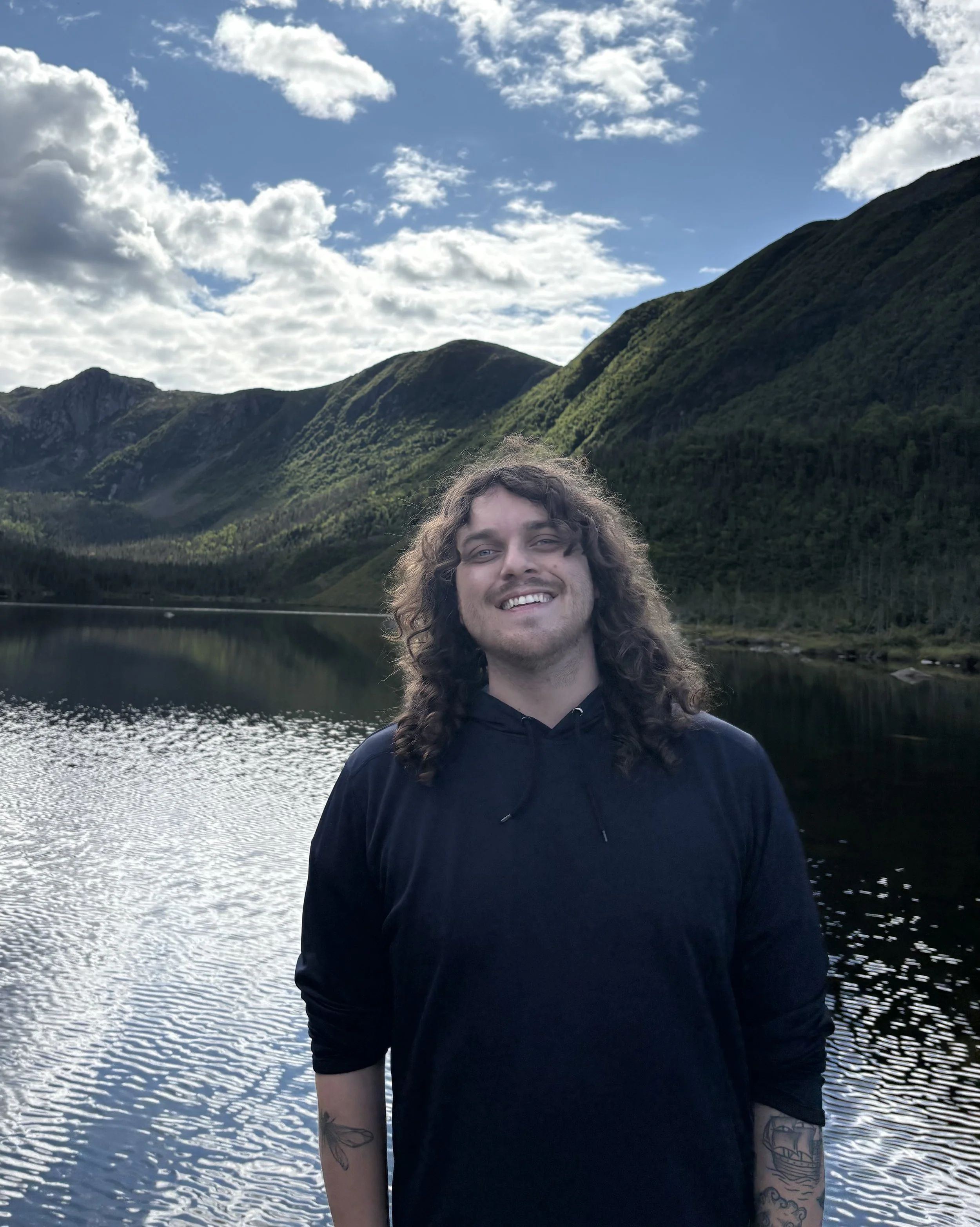 A smiling man with long curly hair and tattoos on his arms, standing in front of a lake with mountains and a partly cloudy sky in the background.