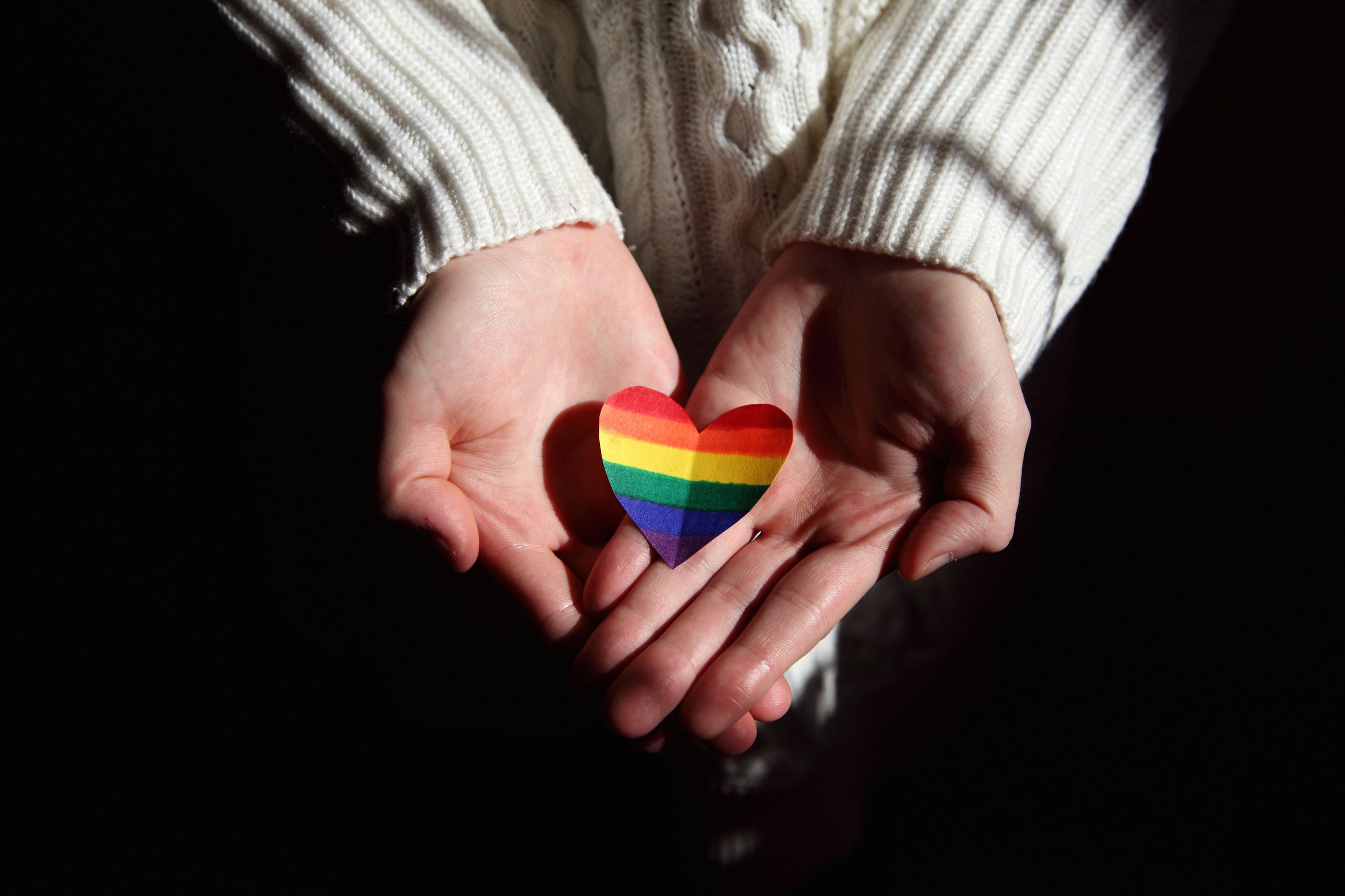 A person holding a paper heart with a rainbow flag on it with a black background