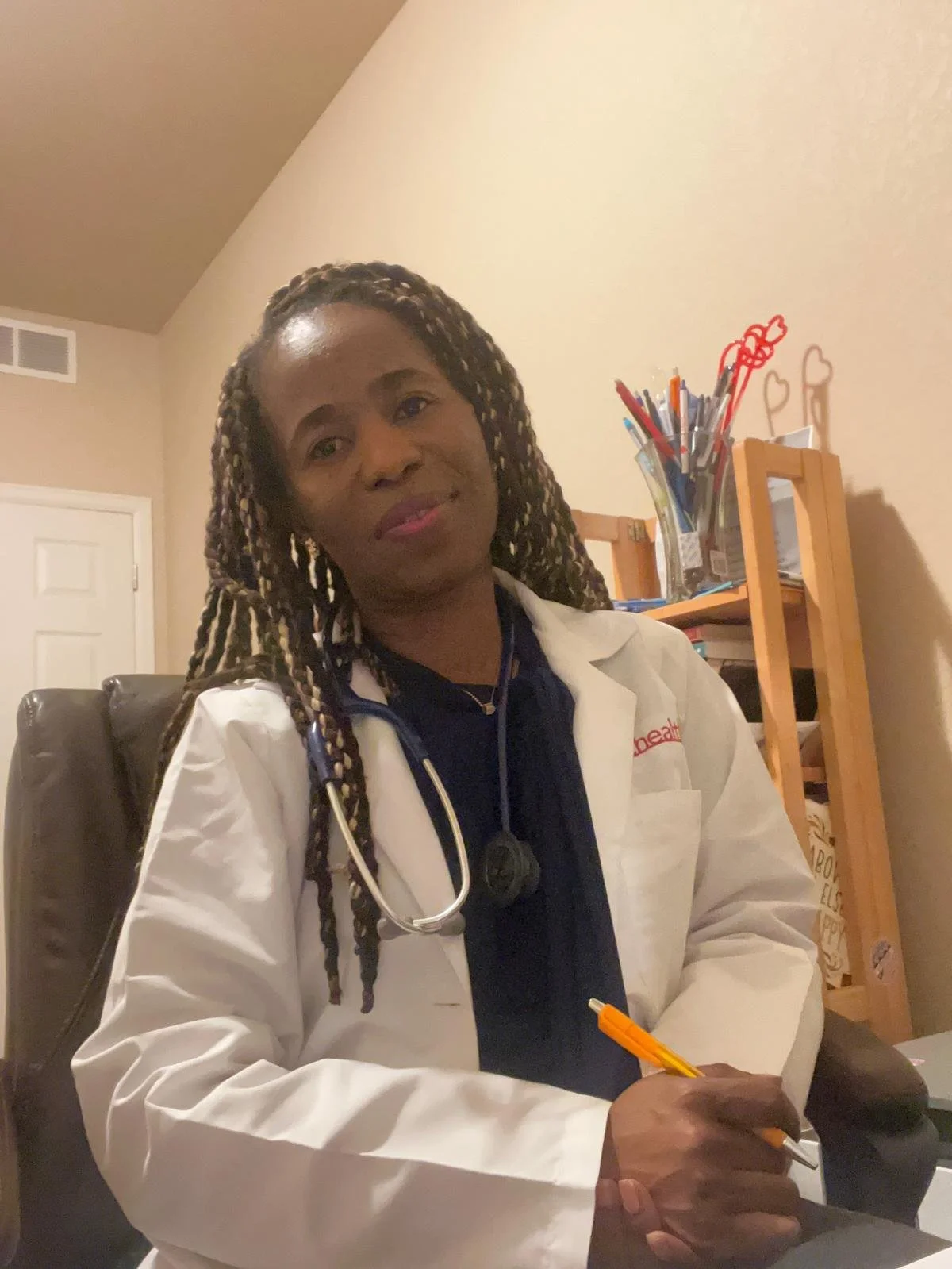 A woman with braided hair sitting at a desk with a stethoscope around her neck, holding a yellow pen, wearing a white coat and dark shirt in an indoor setting with a bookshelf behind her.