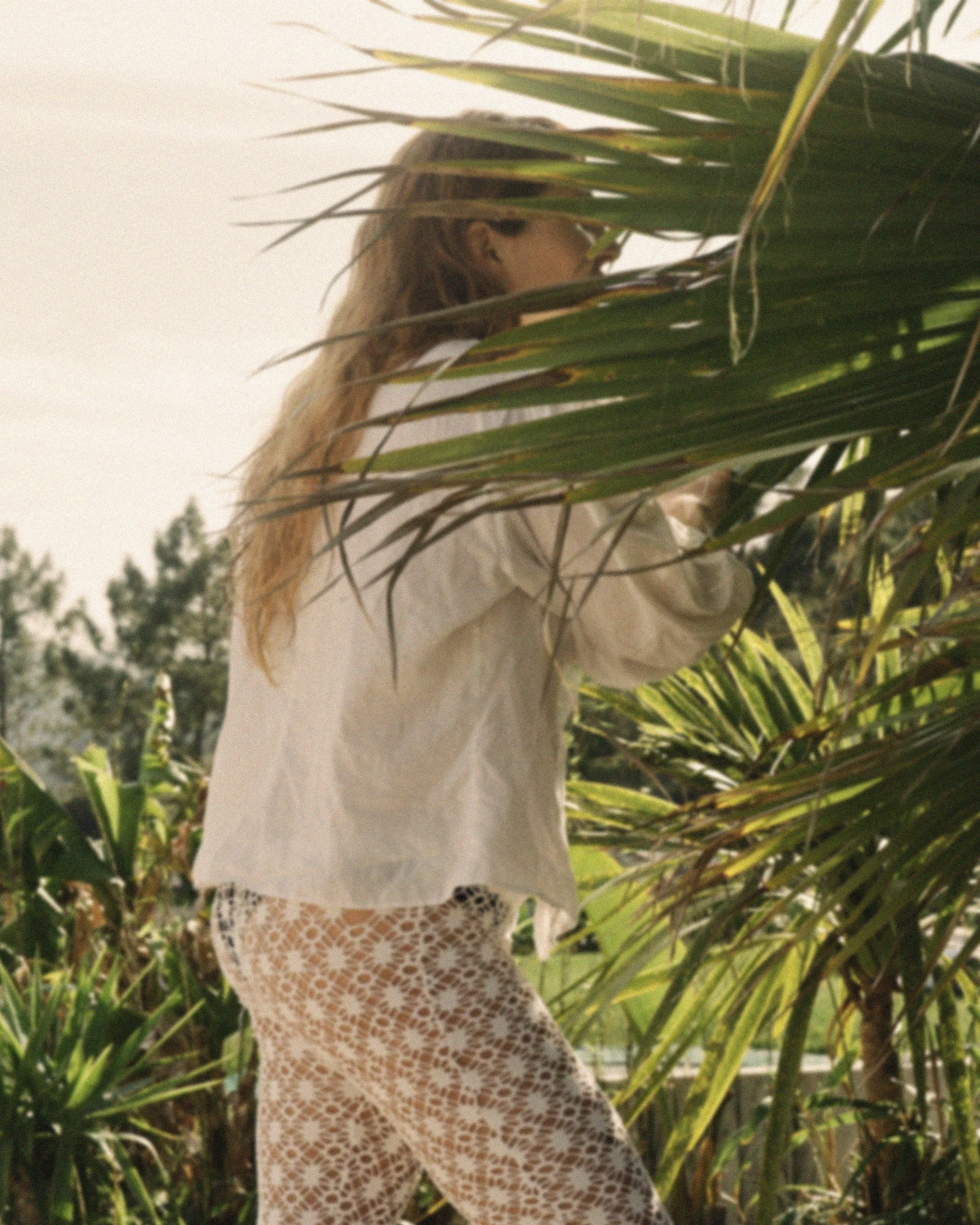 A woman with long hair wearing a white shirt and patterned pants standing among green palm trees.