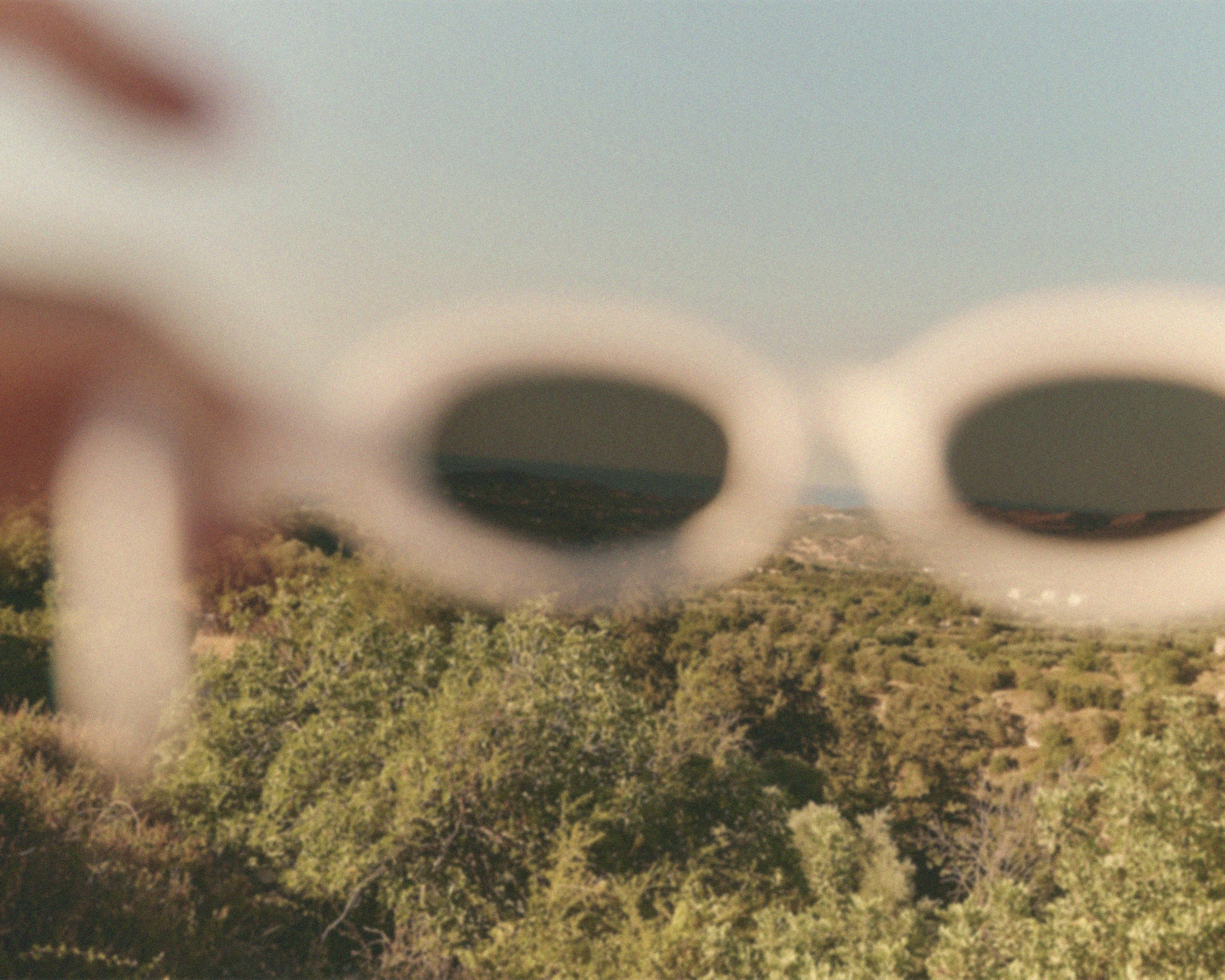 A person holding white sunglasses with round lenses, through which a landscape of trees, hills, and a clear sky is visible.