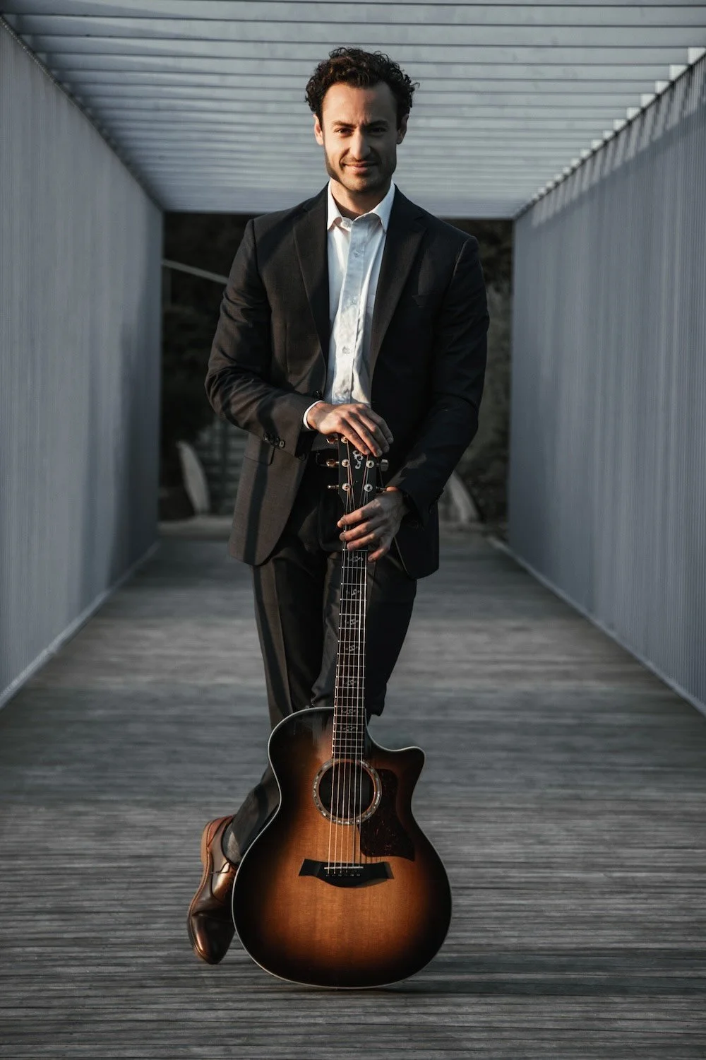 A man in a black suit holding an acoustic guitar on a modern outdoor walkway.