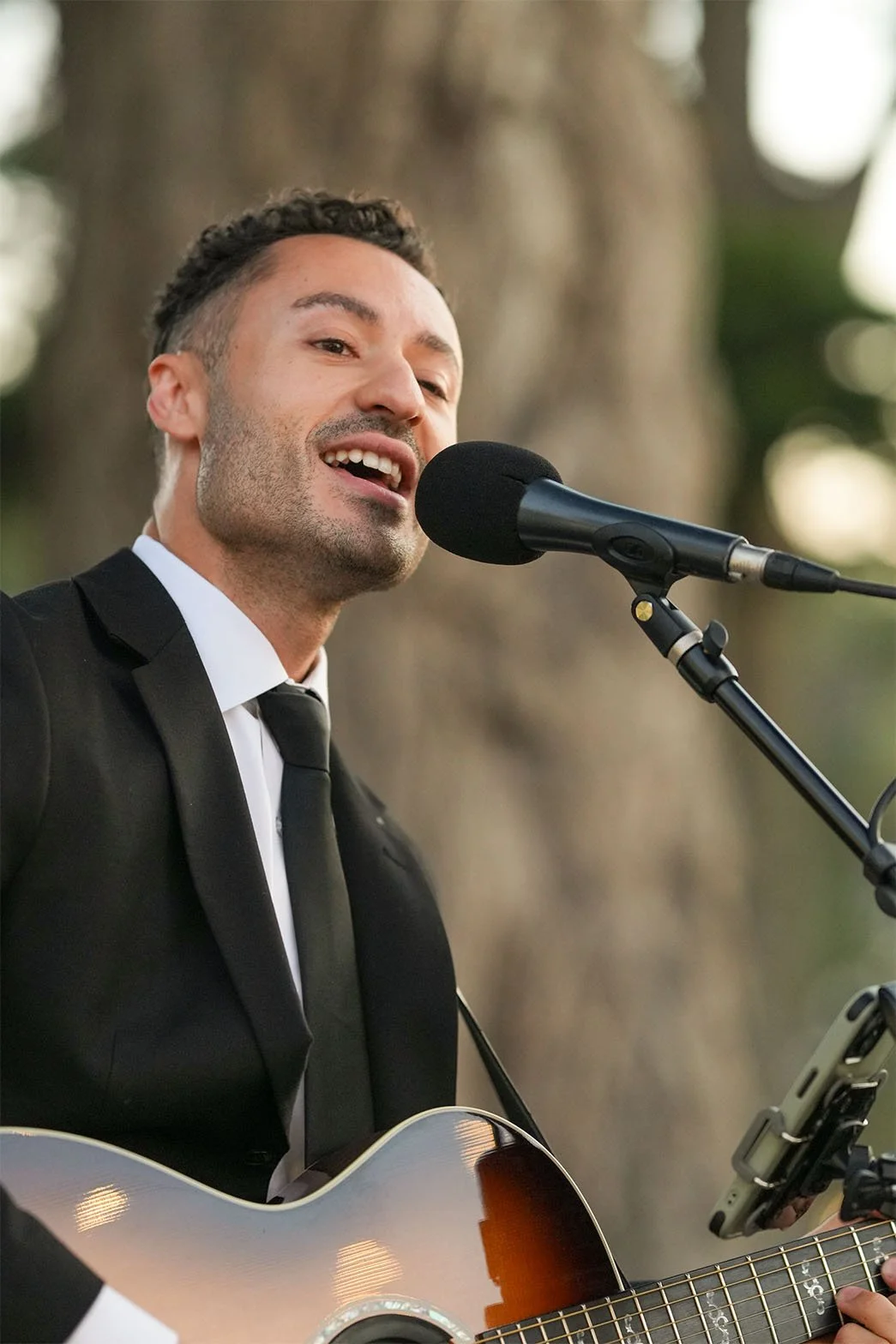 Man in a black suit and white shirt singing into a microphone and playing an acoustic guitar outdoors.