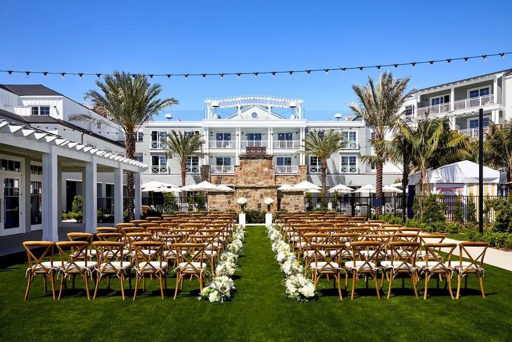 Outdoor wedding ceremony setup with chairs, floral arrangements, white canopy, palm trees, and a large building in the background under a clear blue sky.