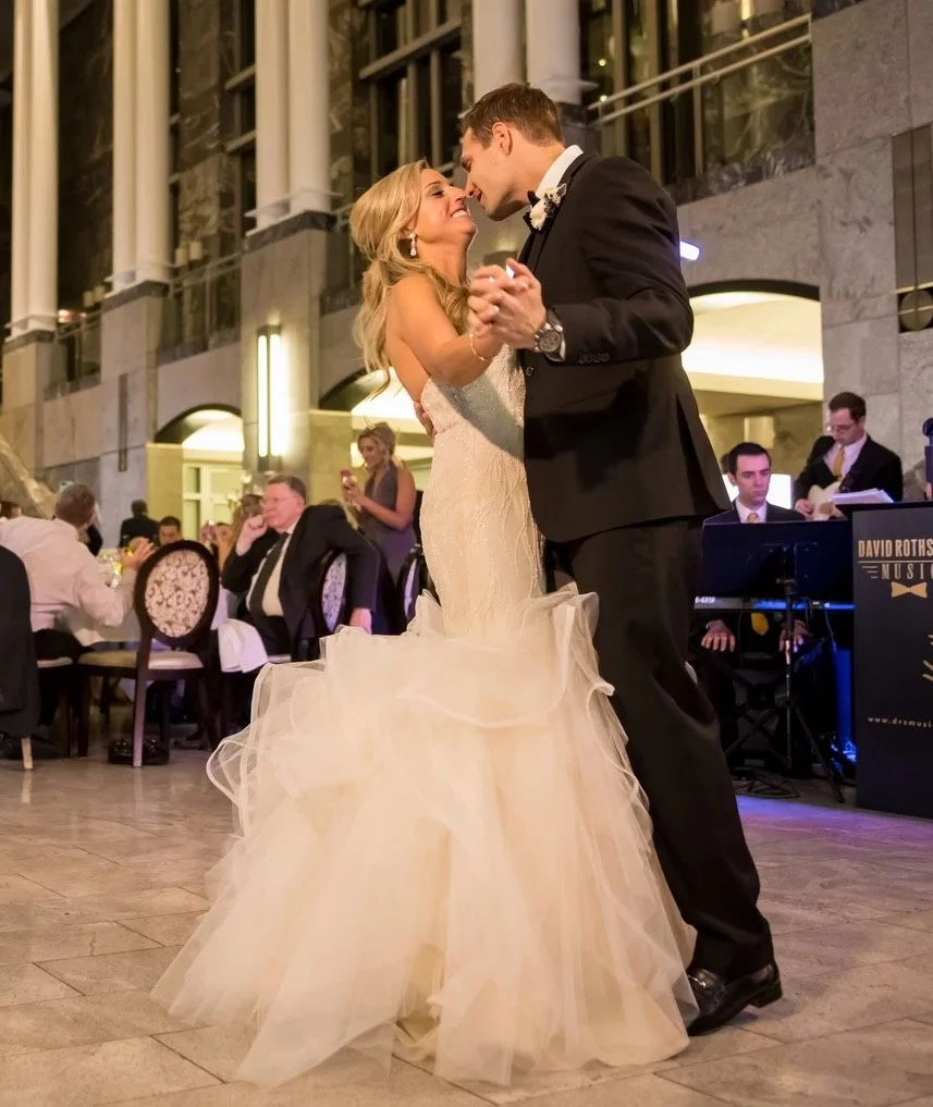 A bride and groom sharing their first dance at a wedding reception in an elegant venue, surrounded by seated guests and a live band.