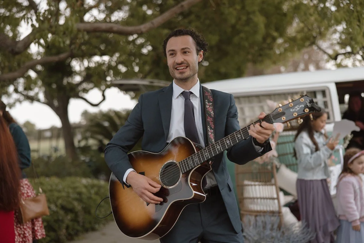 Man in a suit playing an acoustic guitar at an outdoor gathering with children and a van in the background.