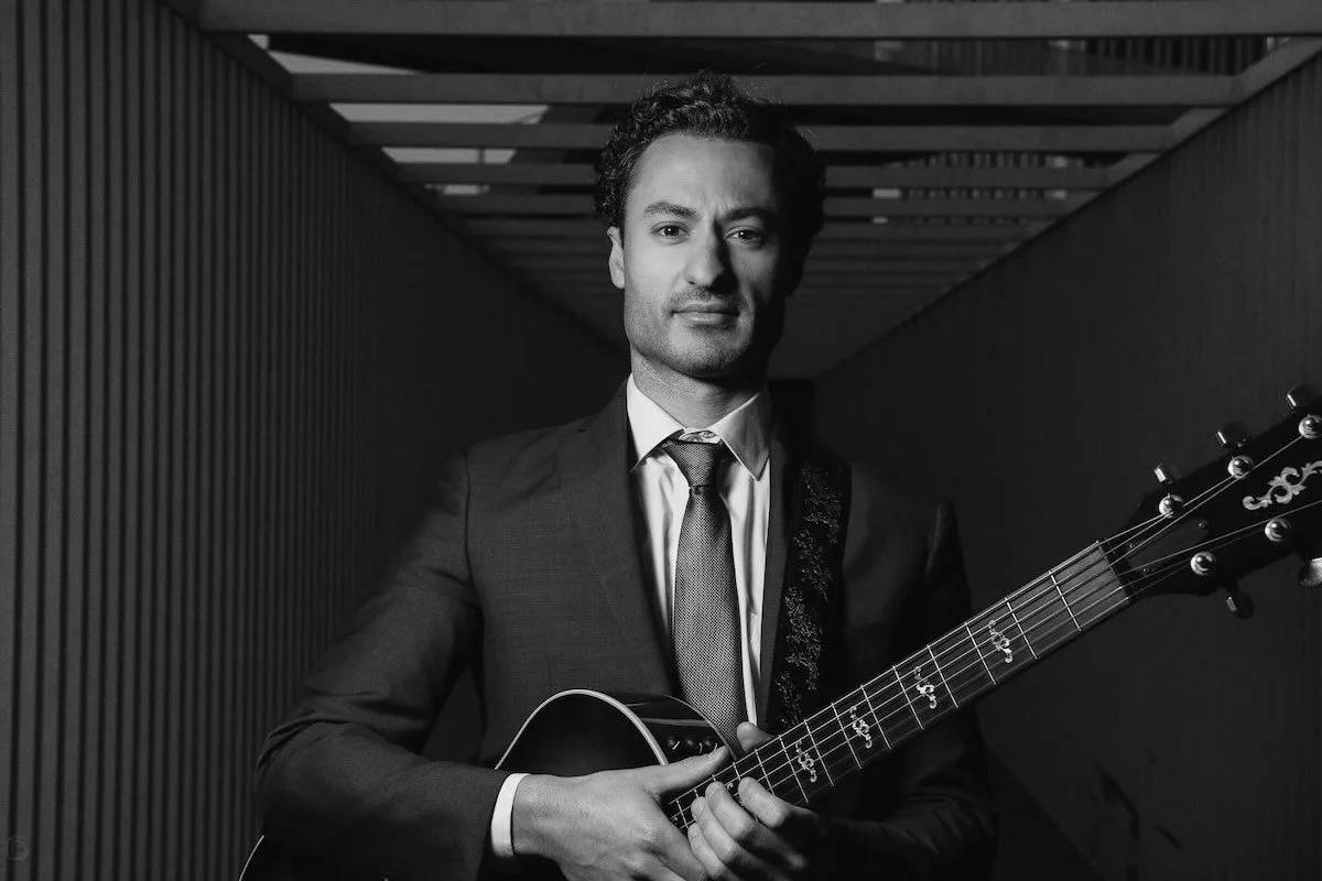 A man in a suit holding an acoustic guitar in a modern, minimalist corridor, black and white photograph.