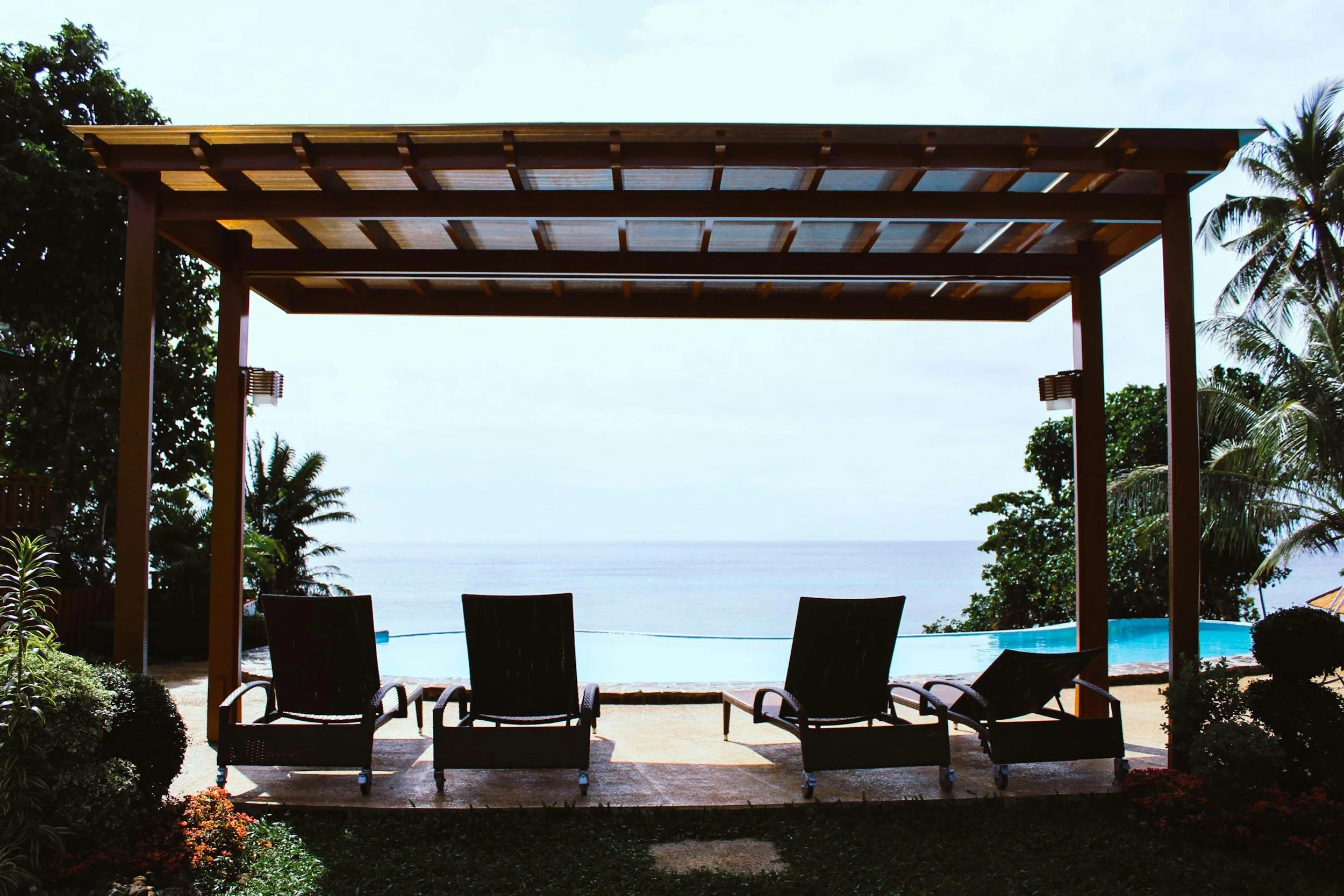 Outdoor pool area with four black lounge chairs and a pool, overlooking the ocean with palm trees and shrubs along the sides, under a wooden pergola.