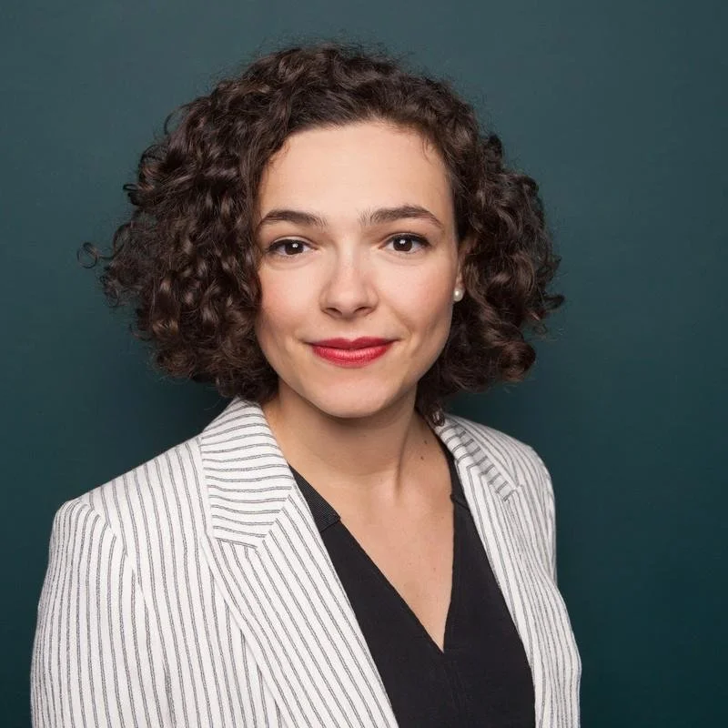 A portrait of a woman with short curly brown hair, red lipstick, wearing a white pinstripe blazer over a black top, standing against a solid dark background.