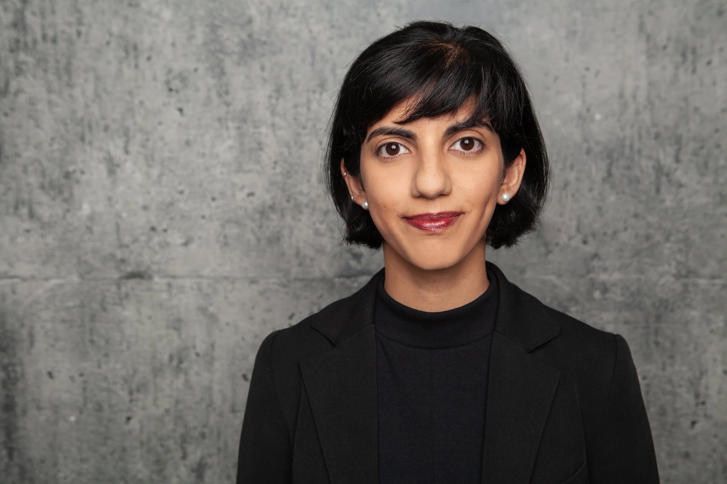 A professional woman with short black hair, wearing a black blazer and pearl earrings, smiling softly against a gray textured background.