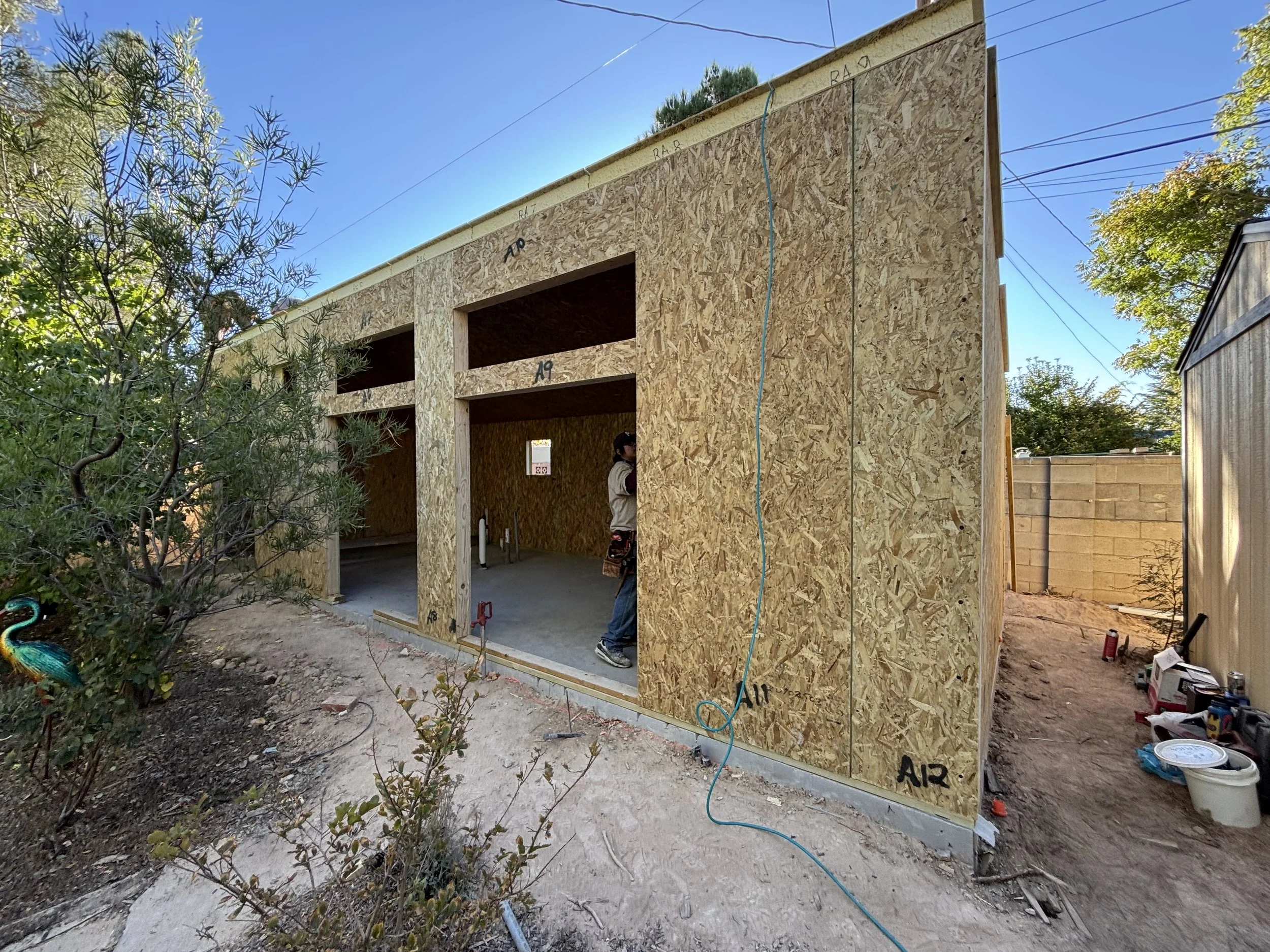 Construction of a small wooden building with OSB walls in progress, with open doorways and a person standing inside, surrounded by construction tools and materials under a clear blue sky.