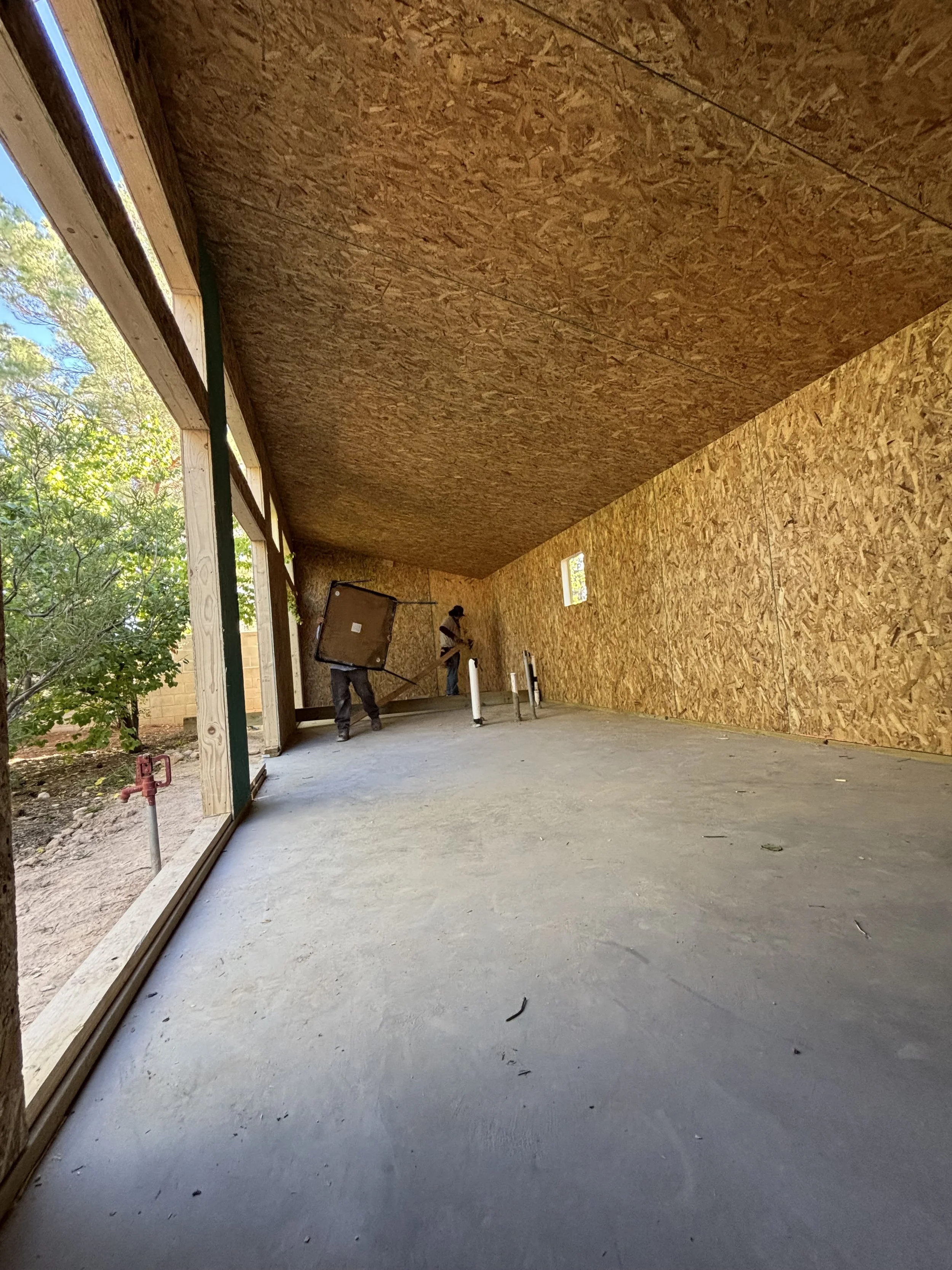 Construction workers inside a partially built wooden structure, with plywood walls and floor, and large open window area showing trees outside.