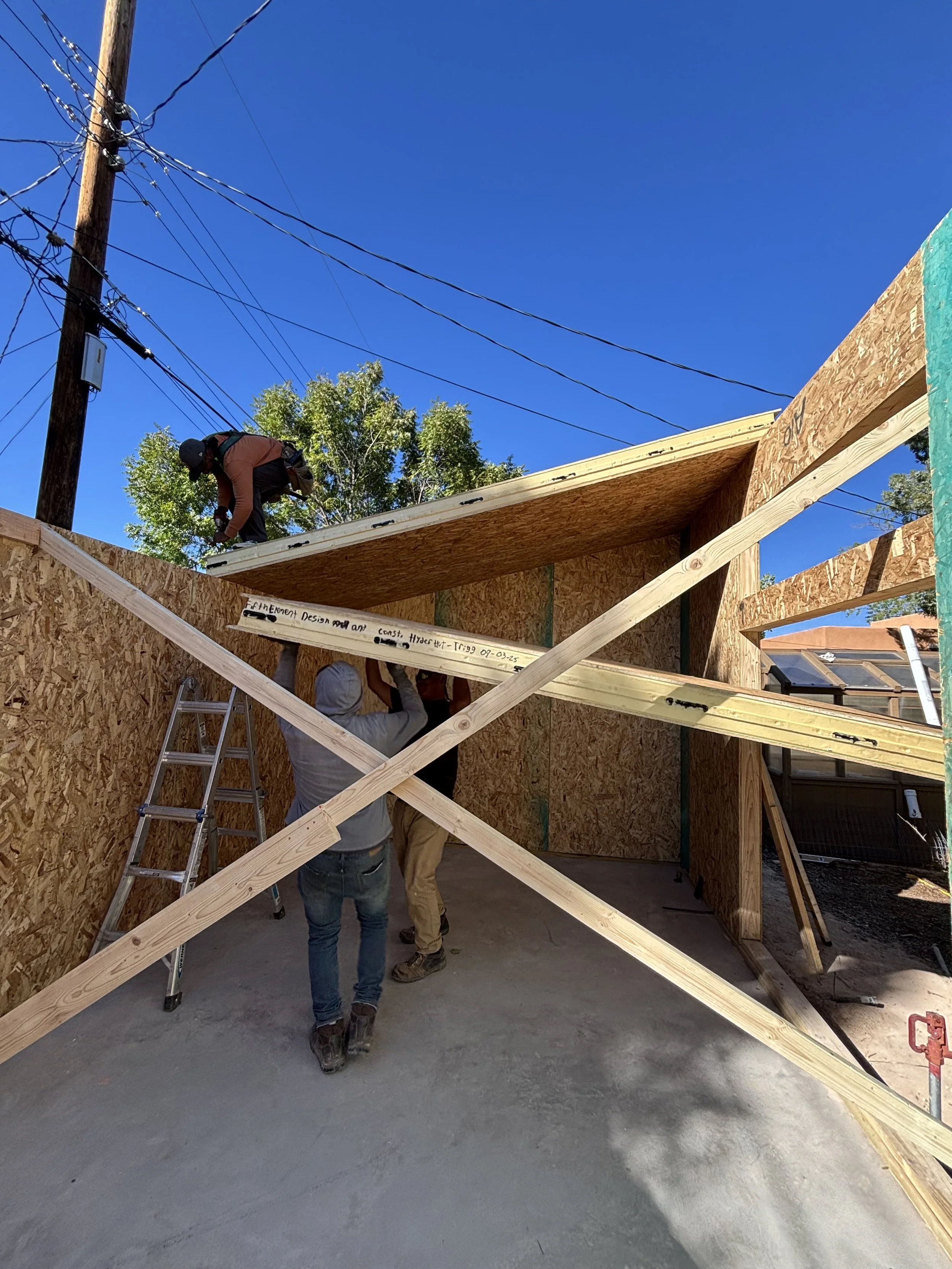 Construction workers building a wooden structure with plywood walls and rafters, under a clear blue sky with overhead power lines and a tree in the background.
