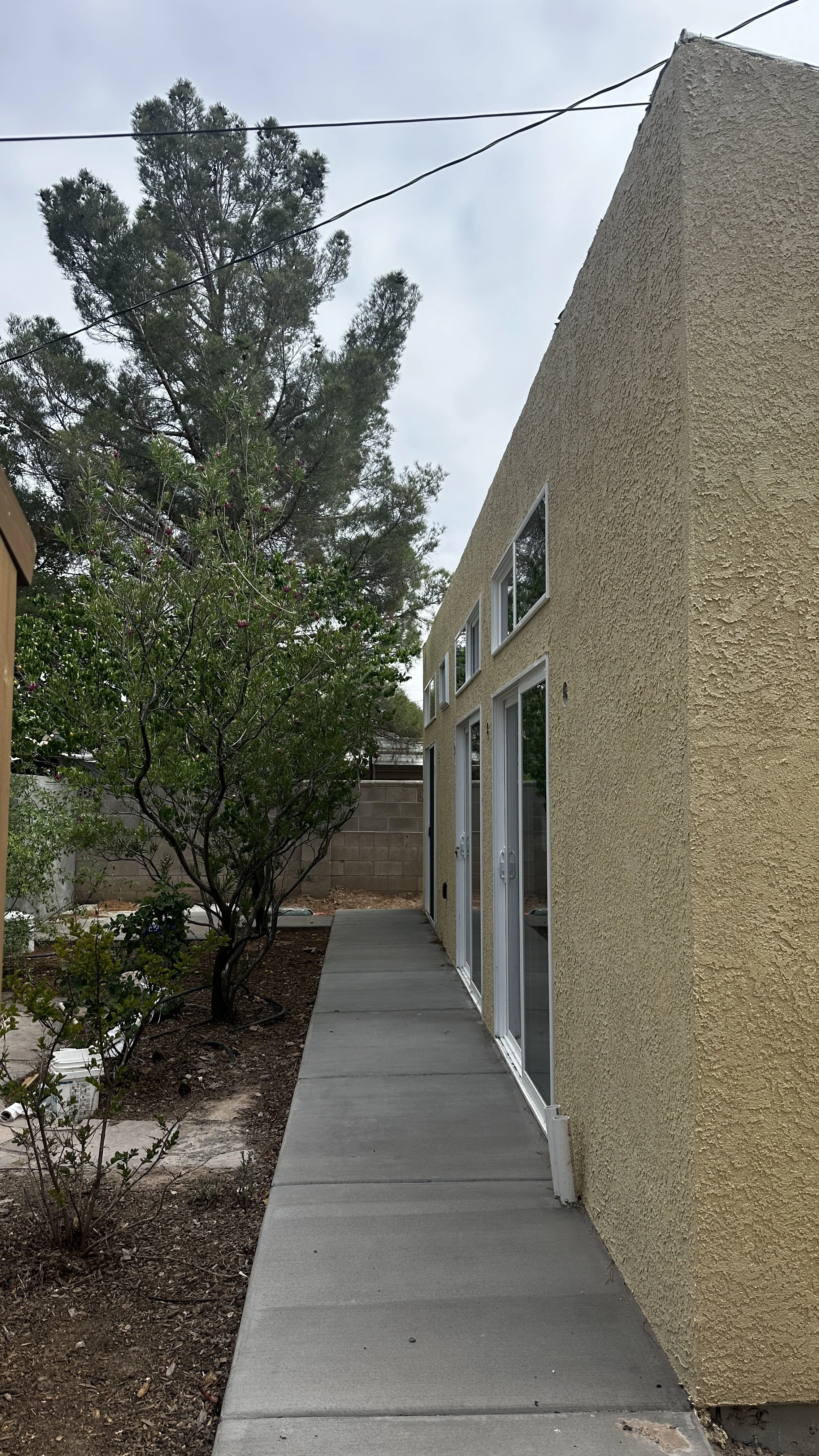 Backyard side of a yellow stucco house with sliding glass doors, a walkway, a small tree, and a cinder block wall in the background.