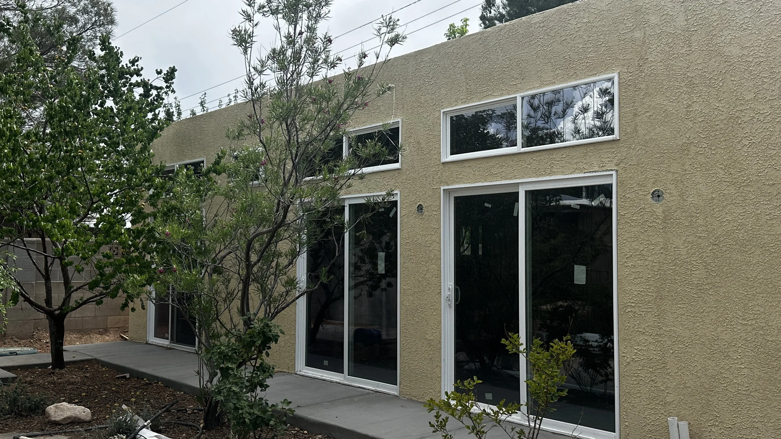 A 420 square foot casita with beige stucco exterior, sliding glass doors, and rectangular windows, surrounded by a yard with trees and plants.