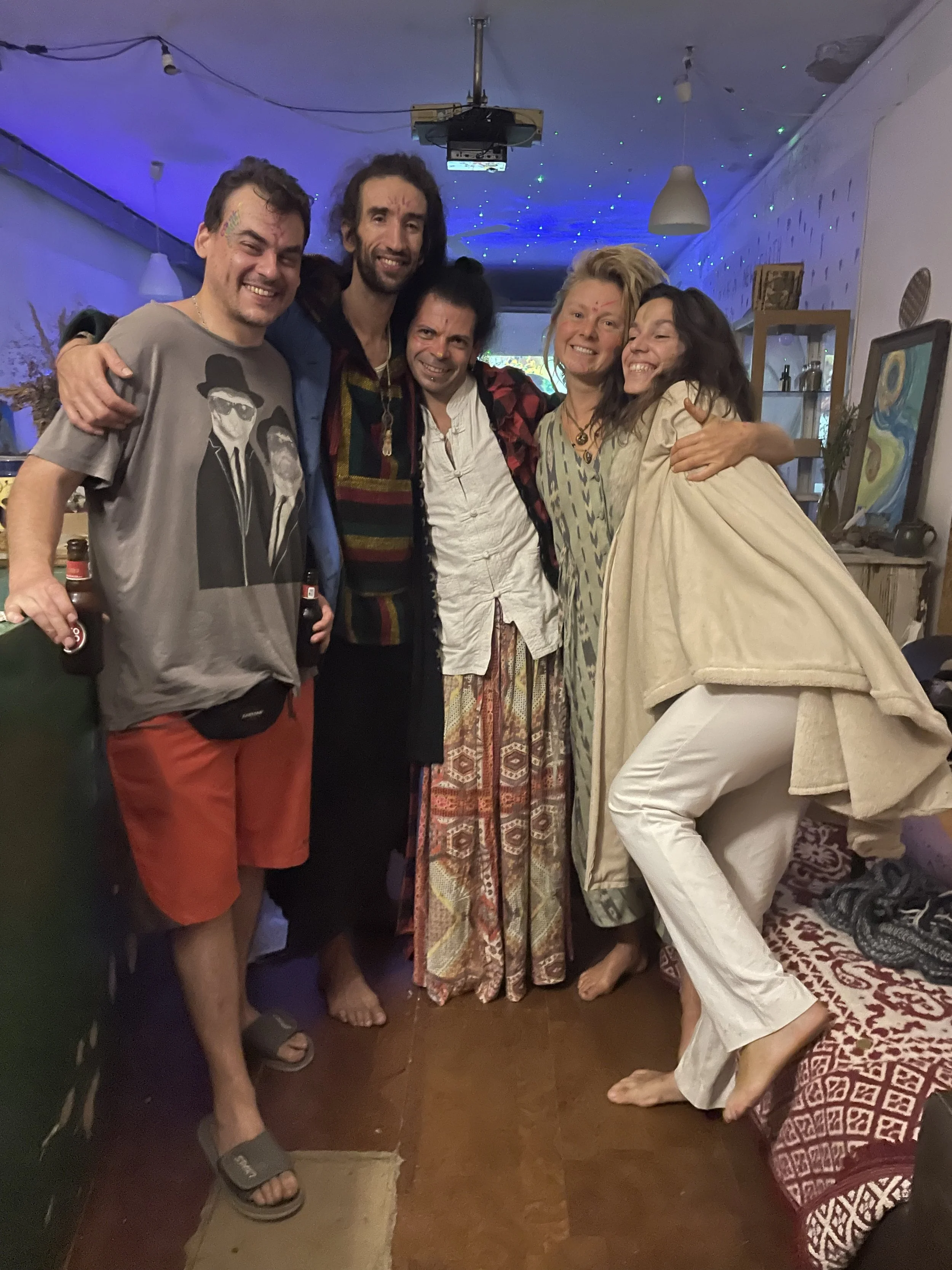 Group of six friends smiling and hugging indoors, with colorful lights and cozy decor in the background.