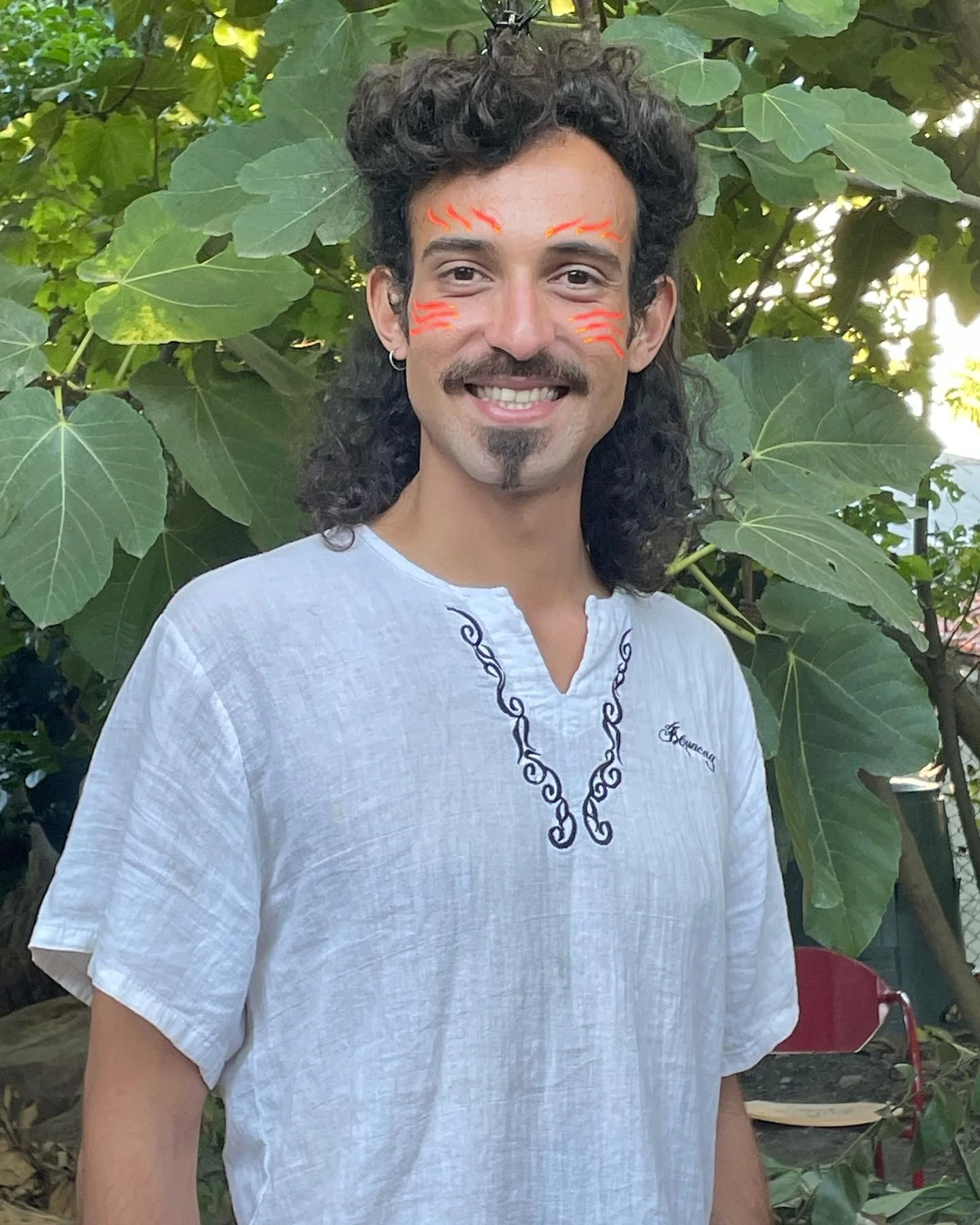 A smiling man with curly hair and a goatee, decorated with orange face paint resembling flames on his forehead and cheeks, standing outdoors in front of large green leaves.