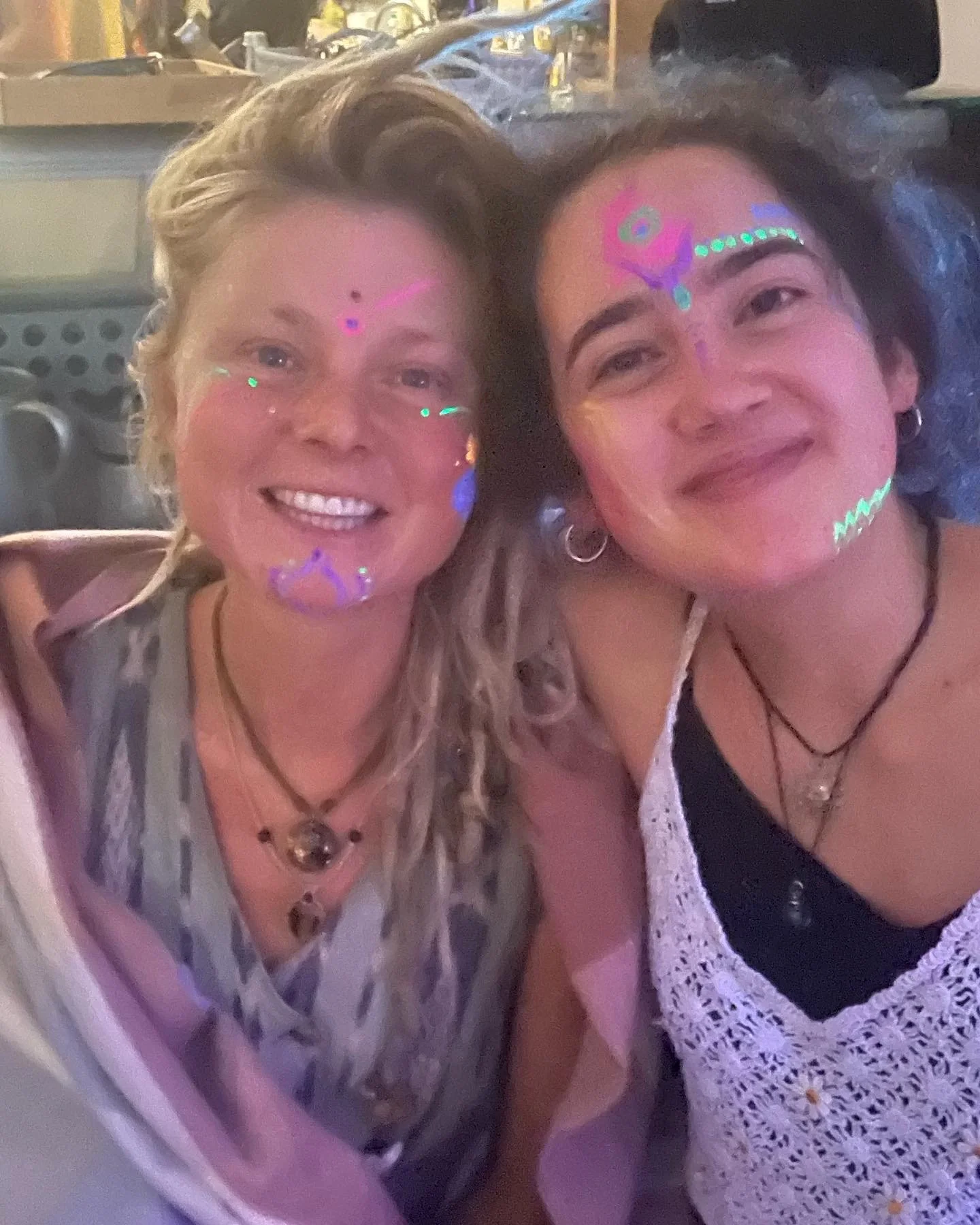 Two women smiling at a party with colorful face paint and glow-in-the-dark markings. One has light skin and blonde hair, the other has darker skin and dark curly hair.