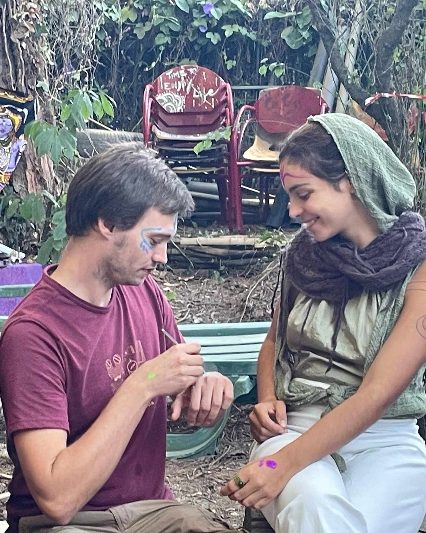 A man and woman sitting outdoors, getting henna tattoos on their hands and faces, with chairs stacked in the background and surrounded by greenery.