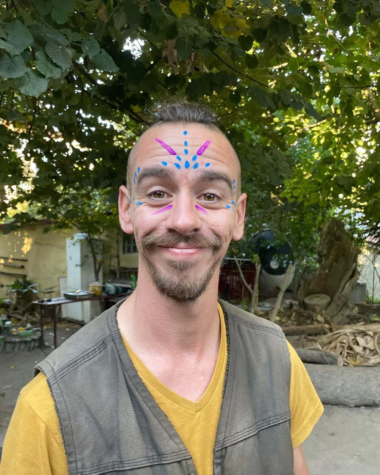 A smiling man with face paint in blue and pink, standing outdoors under trees.