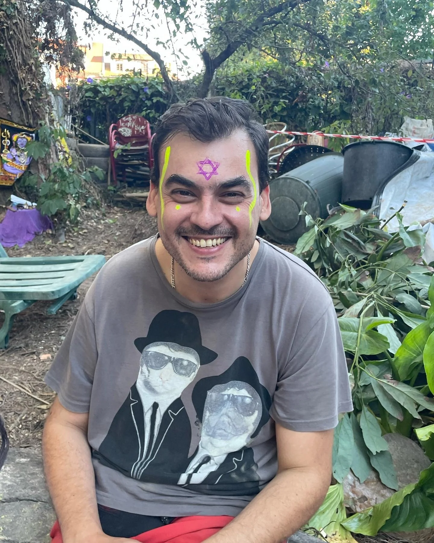 A smiling man with face paint, including a purple Star of David on his forehead and yellow lines on his cheeks, sitting outdoors surrounded by plants, garden tools, and stacked chairs.