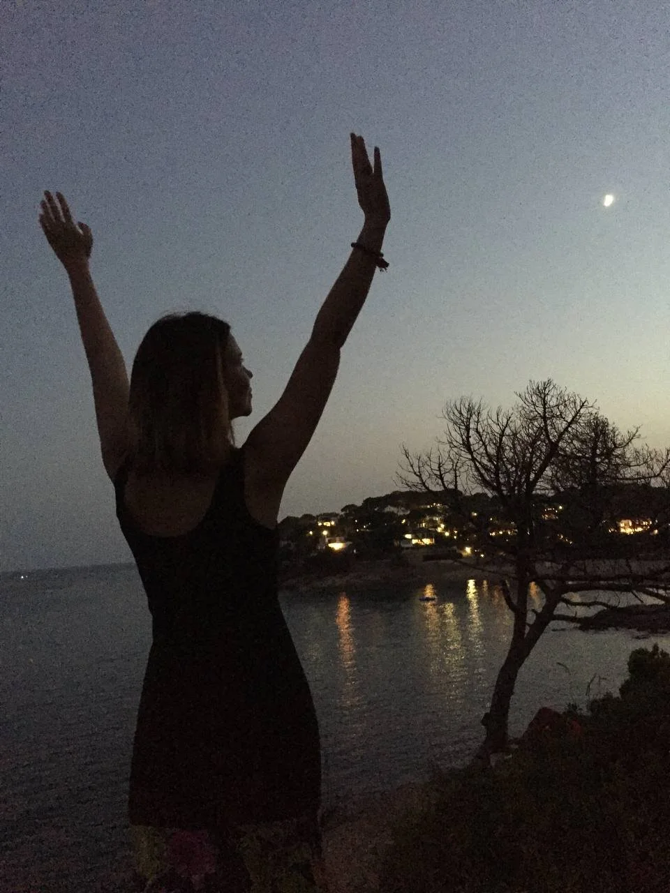 A woman standing outdoors at dusk with her arms raised, silhouetted against a darkening sky, near water and a tree with scattered lights in the background.