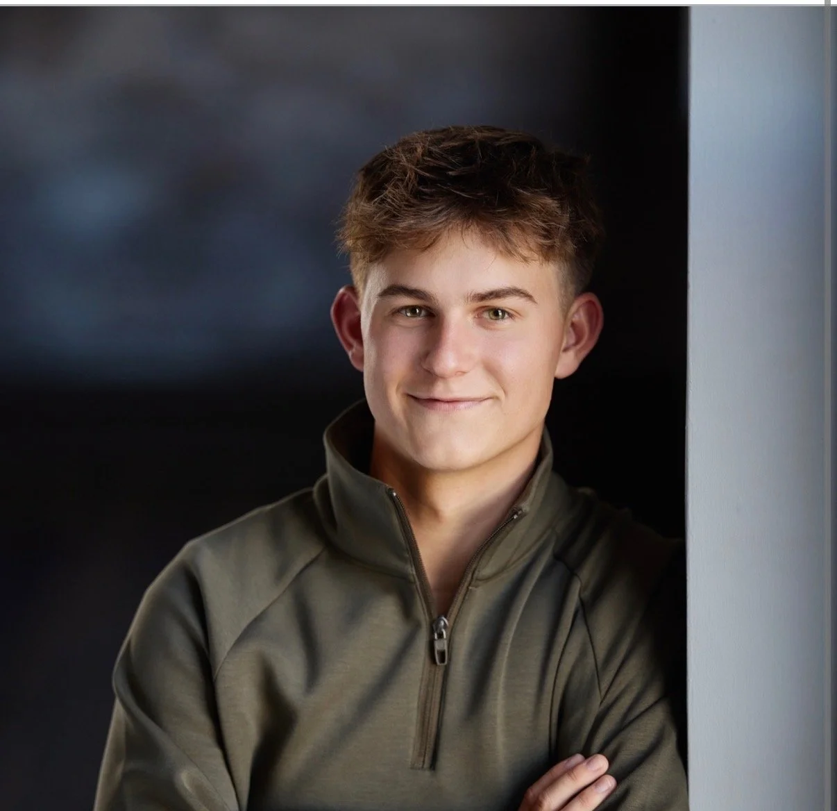 A young man with short, light brown hair smiling, wearing a zip-up jacket, standing with his arms crossed against a dark background.