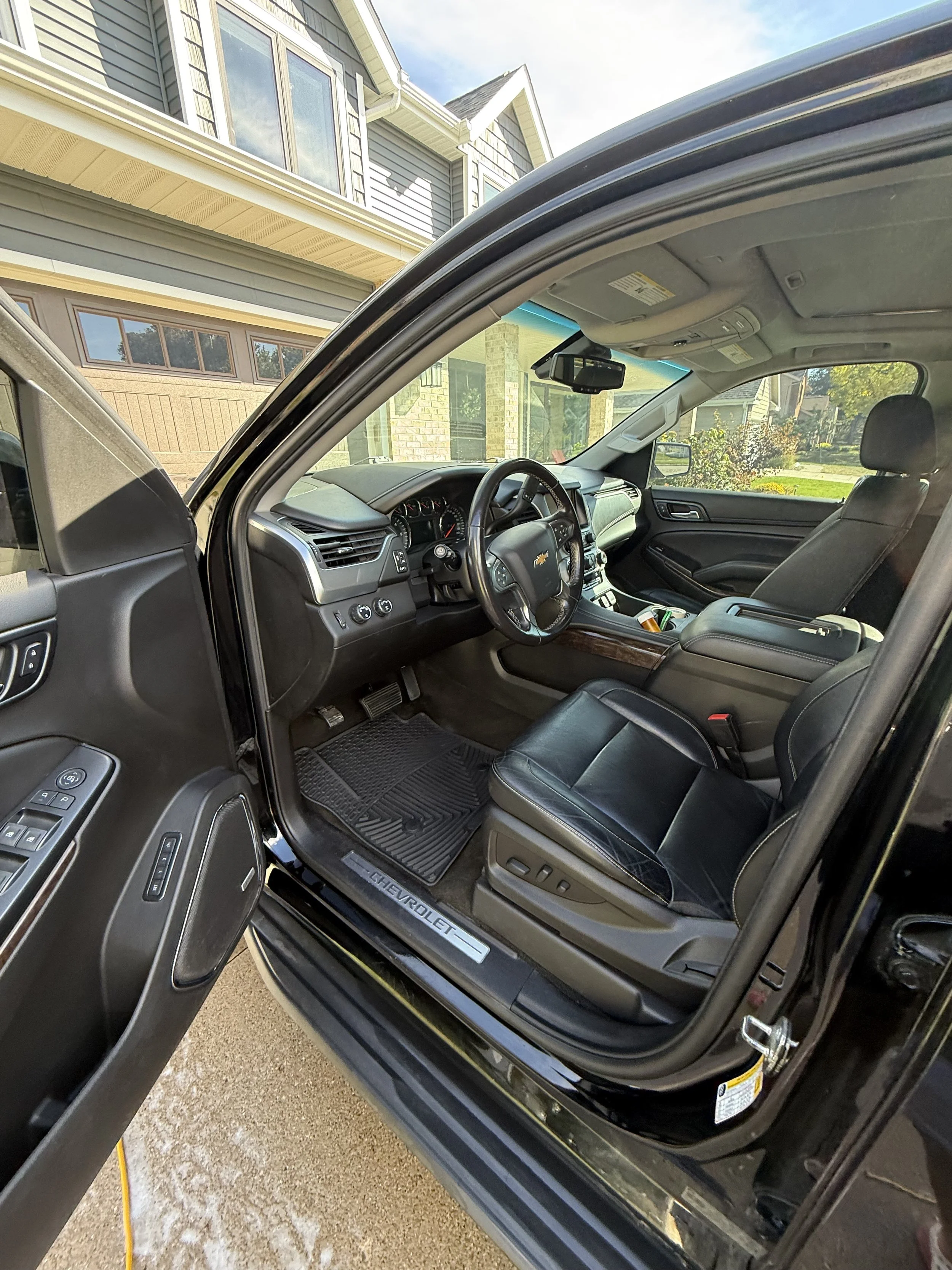 Inside view of a black Chevrolet vehicle with the driver's door open, showing the black leather seats, dashboard, steering wheel, and some controls, parked outside a house.