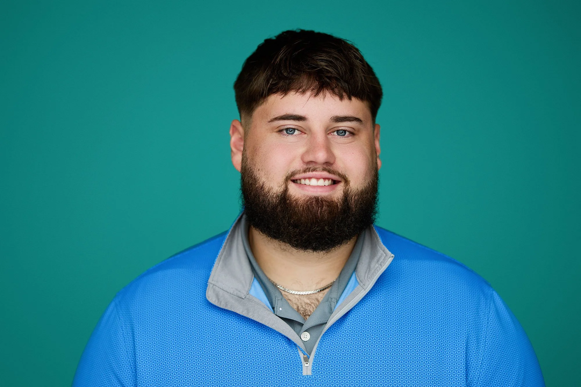 Portrait of a young man with a beard and short dark hair, smiling, wearing a blue zip-up athletic jacket over a gray polo shirt, against a solid teal background.