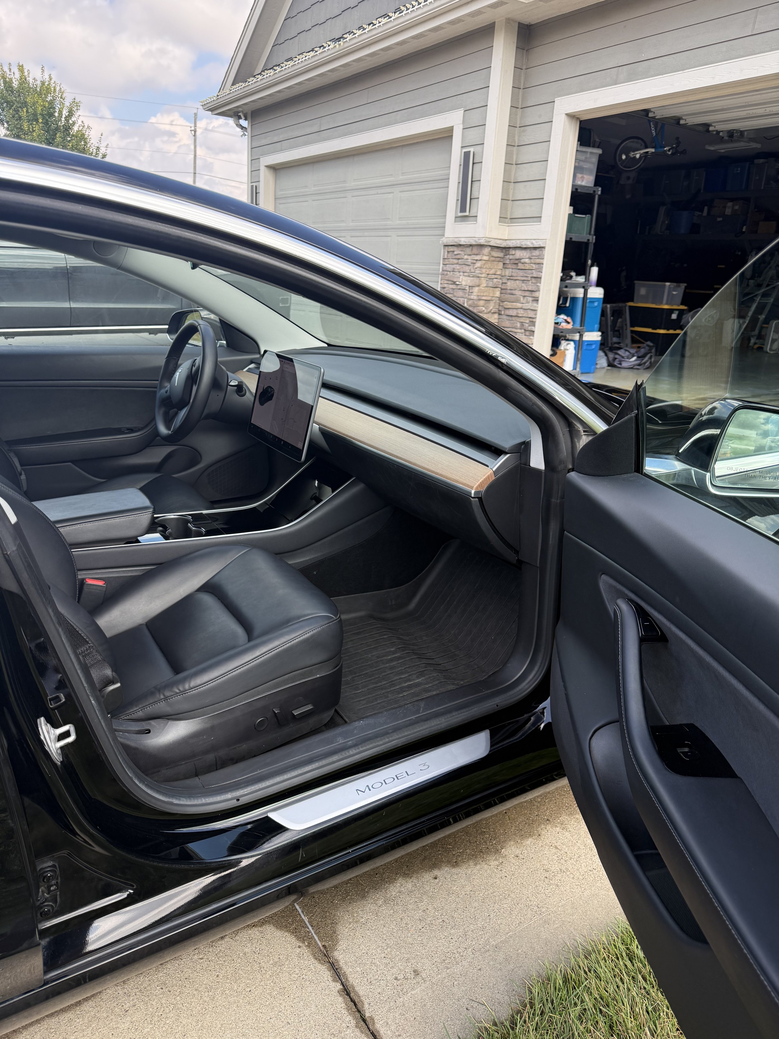 Open black Tesla Model 3 showing the interior with black leather seats, a central touchscreen display, and a dashboard with wood accent. The car is parked outside a house with a garage in the background.