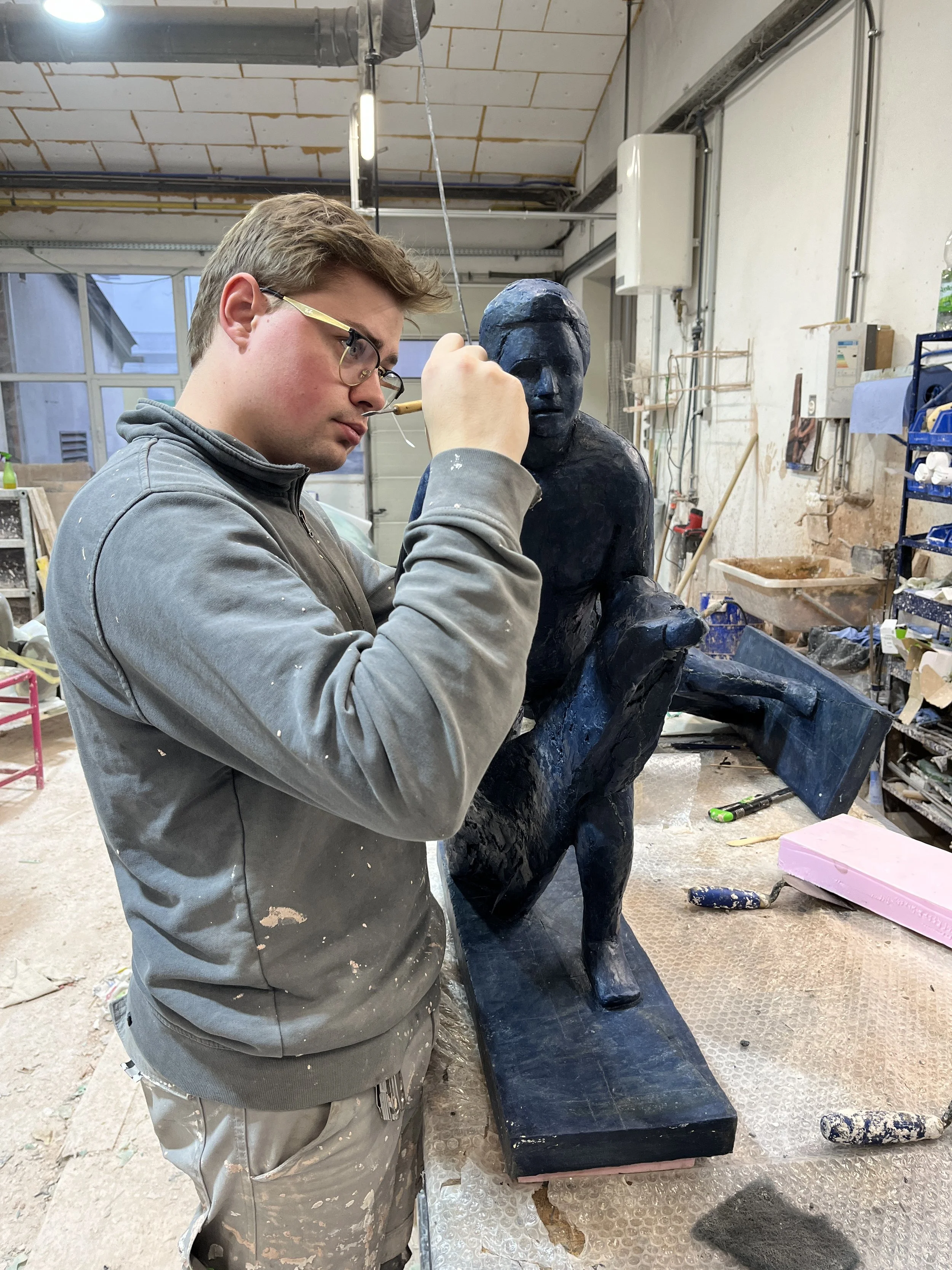 A person wearing glasses and a gray hoodie works on the wax model for a metal sculpture of a seated human figure in an indoor art studio.
