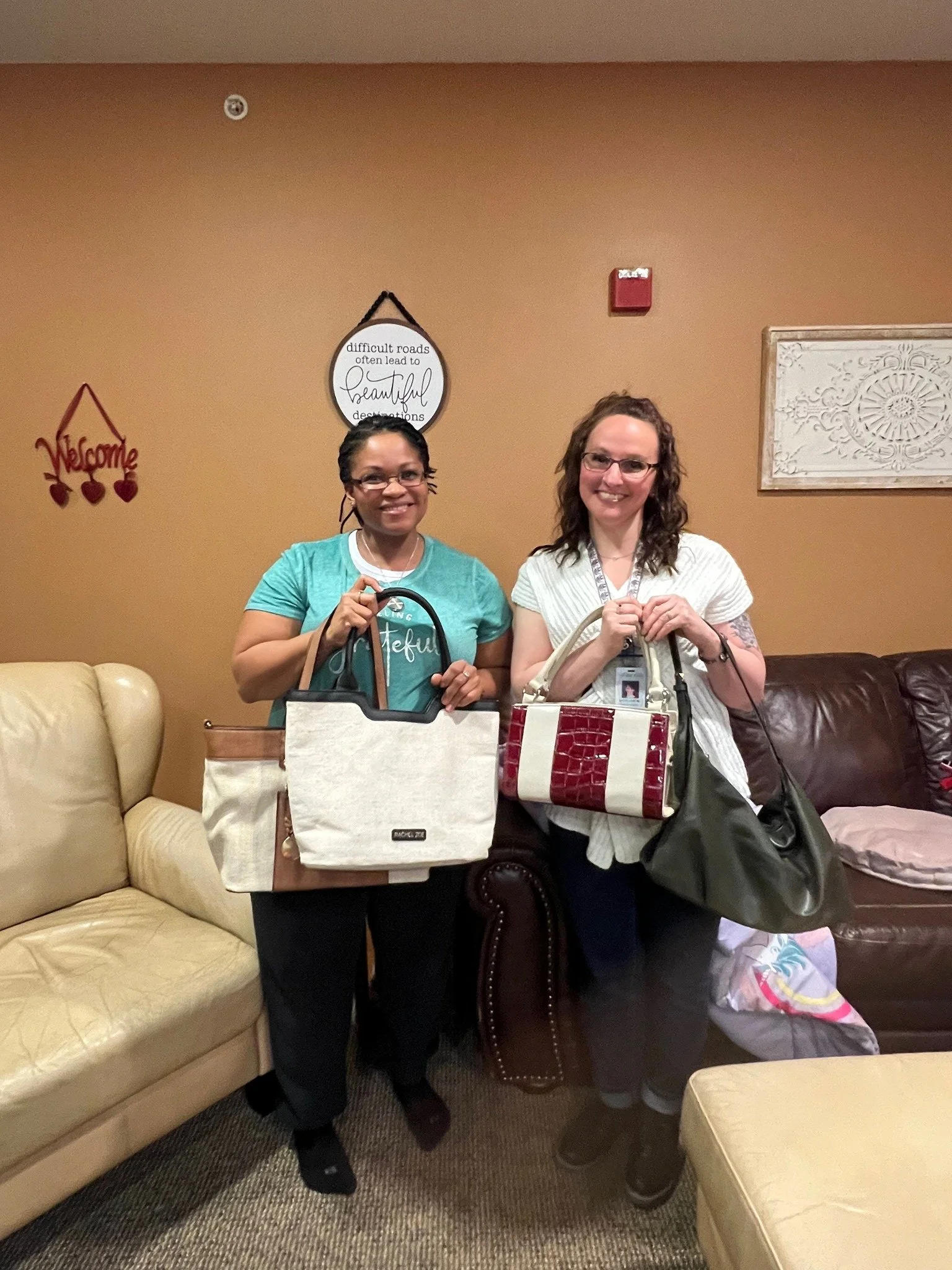 Two women standing side by side in a living room holding handbags, with beige and brown couches and decorative wall signs behind them.