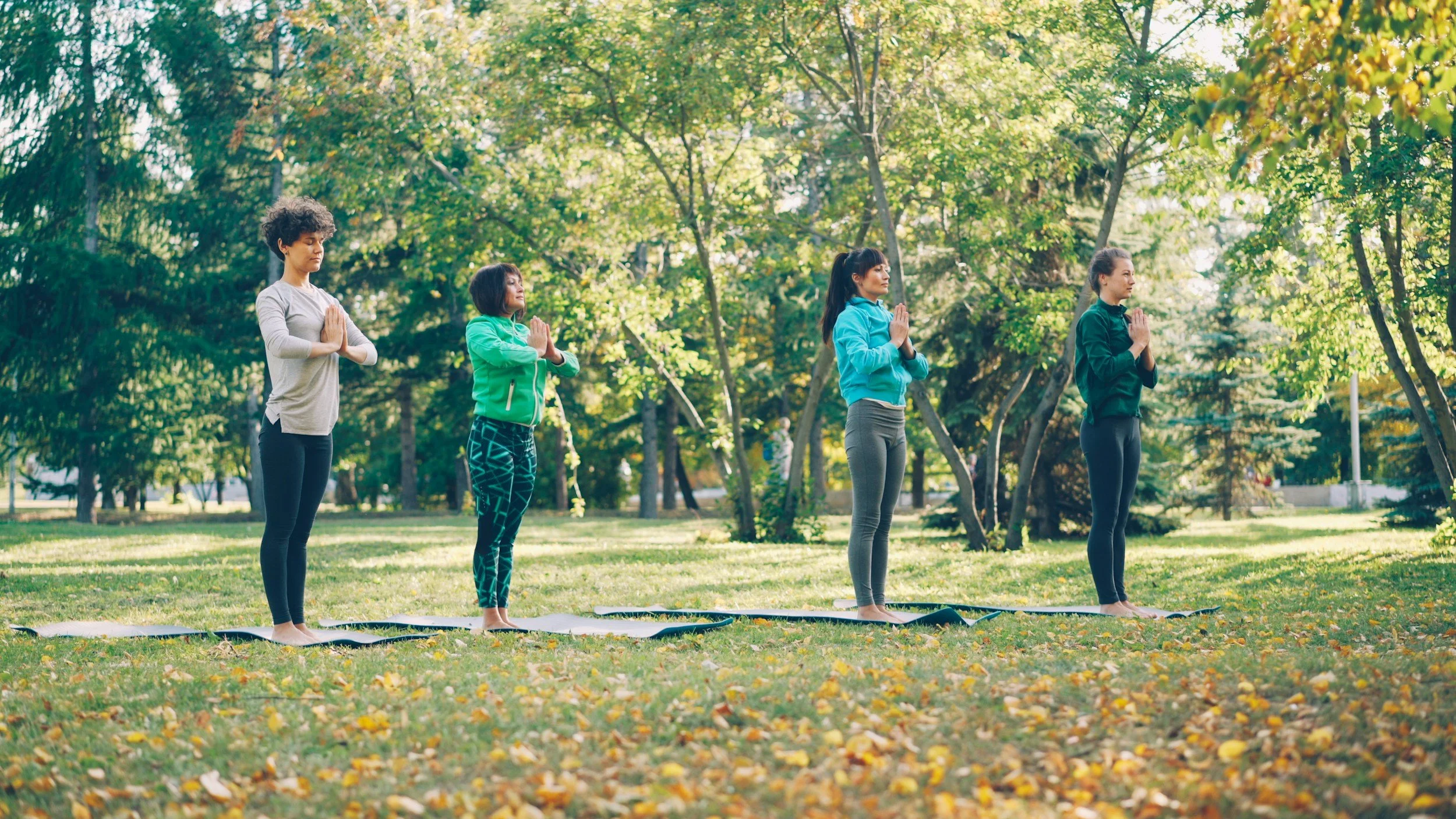 Four women practicing yoga outdoors on mats in a park with green trees and fallen leaves.