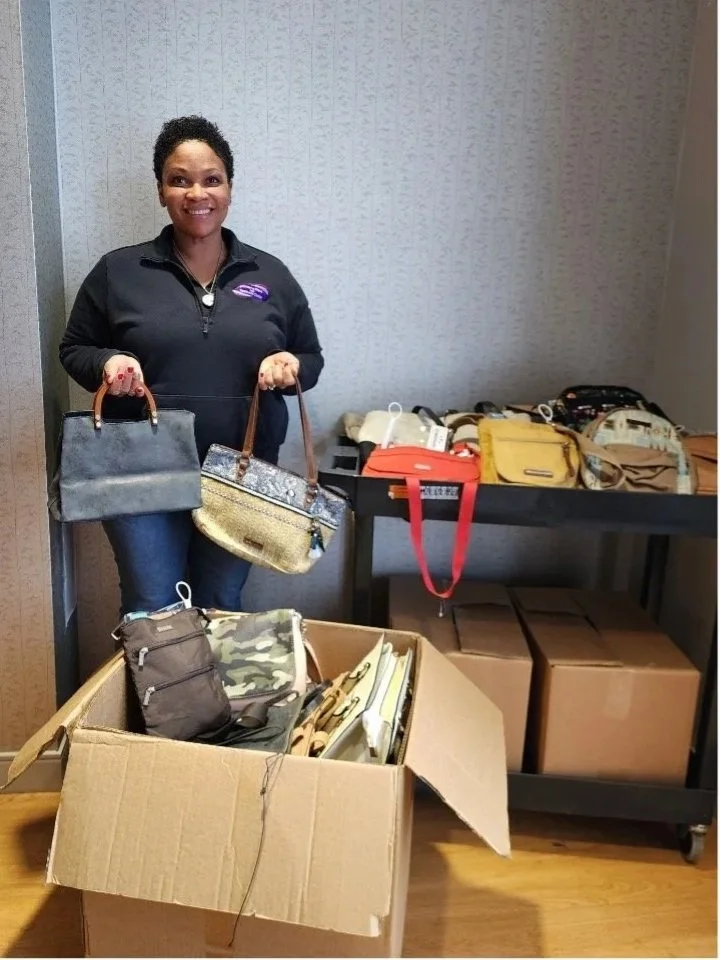 Woman smiling and holding two handbags, standing next to a table with bags and a box filled with miscellaneous items.