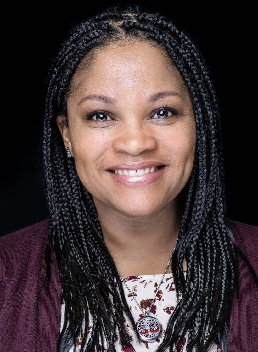 A woman with braided hair smiling, wearing earrings, a floral blouse, and a purple blazer with a circular pendant necklace, against a black background.