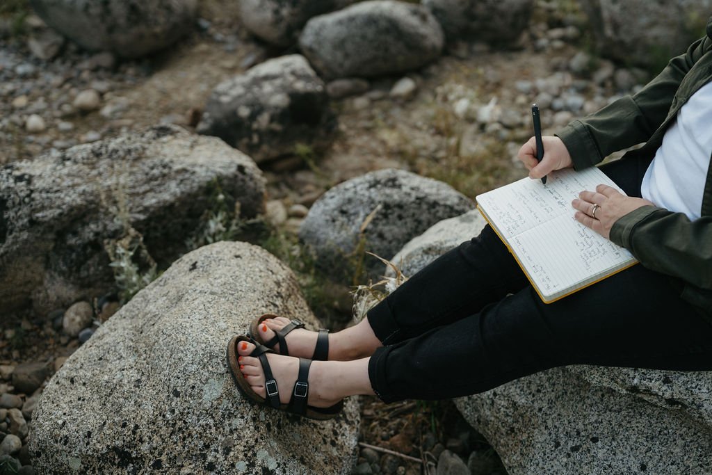 Feet in open toed sandals writing in a journal while sitting on rocks.