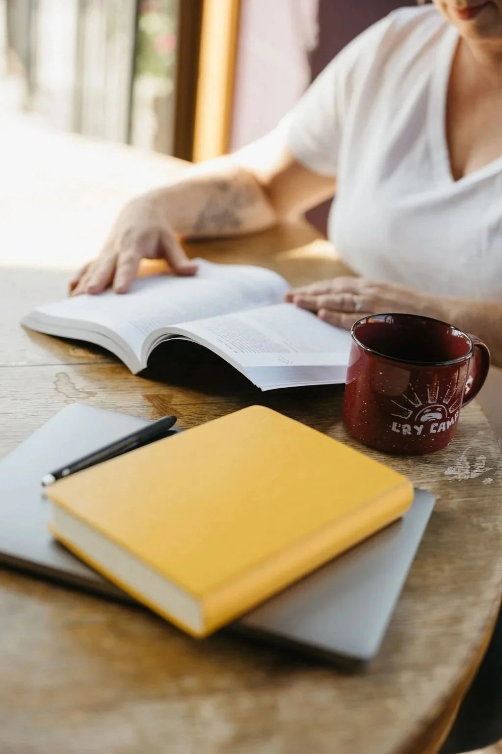 Yellow journal sitting on top of a laptop while a woman read as book drinking coffee out of a red mug